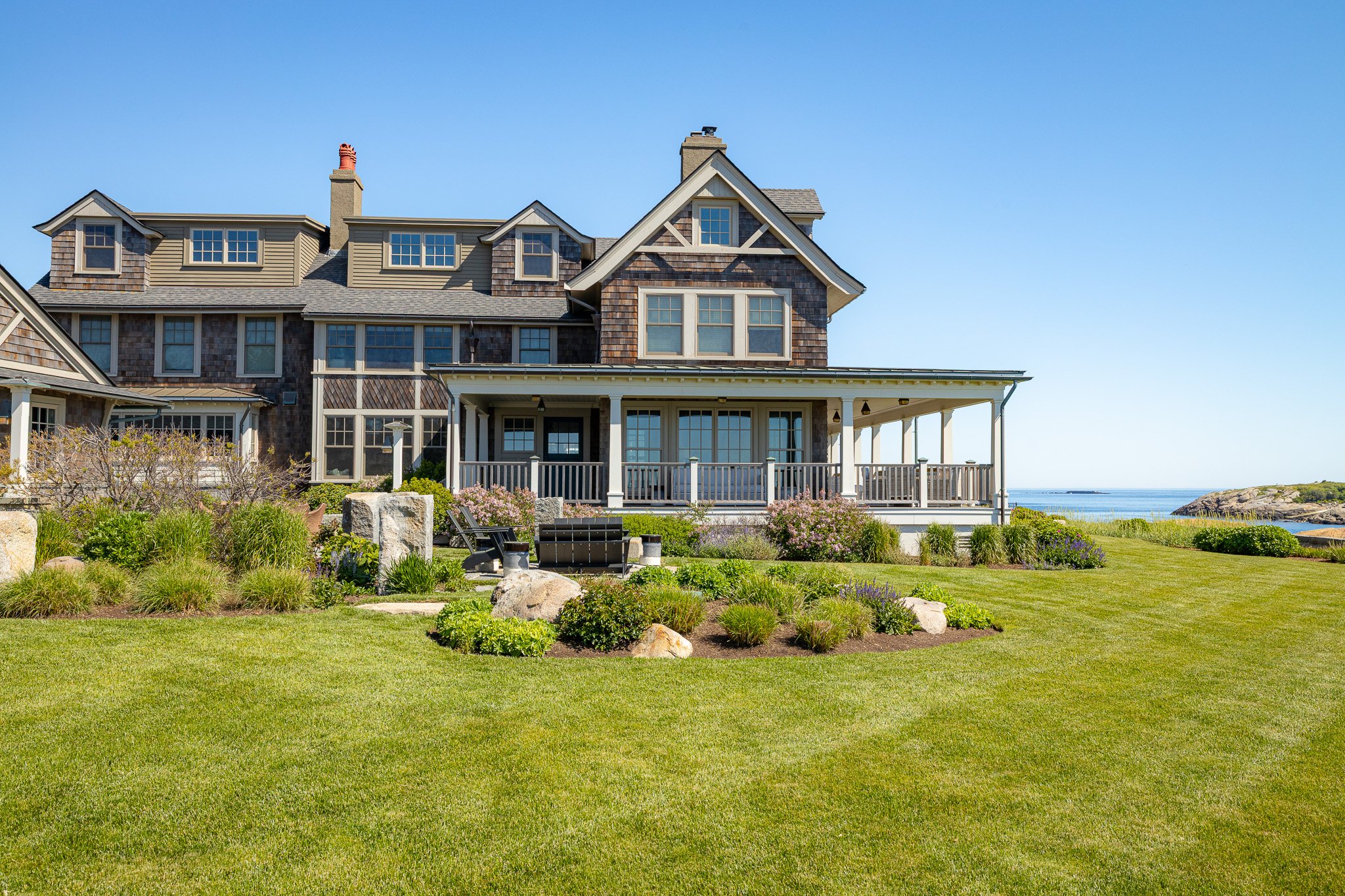 Large oceanfront house with multiple stories, gabled roof, and expansive porch, surrounded by landscaped lawn, flowers, rocks, and overlooking water.