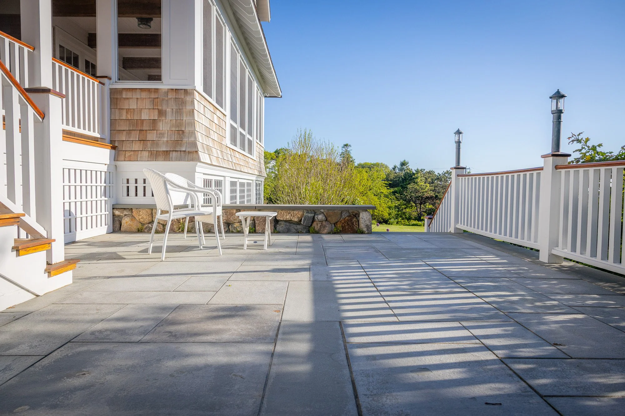A spacious outdoor patio with large gray tiles, white chairs, and a low stone wall, surrounded by white wooden fences and lush green trees under a clear blue sky.