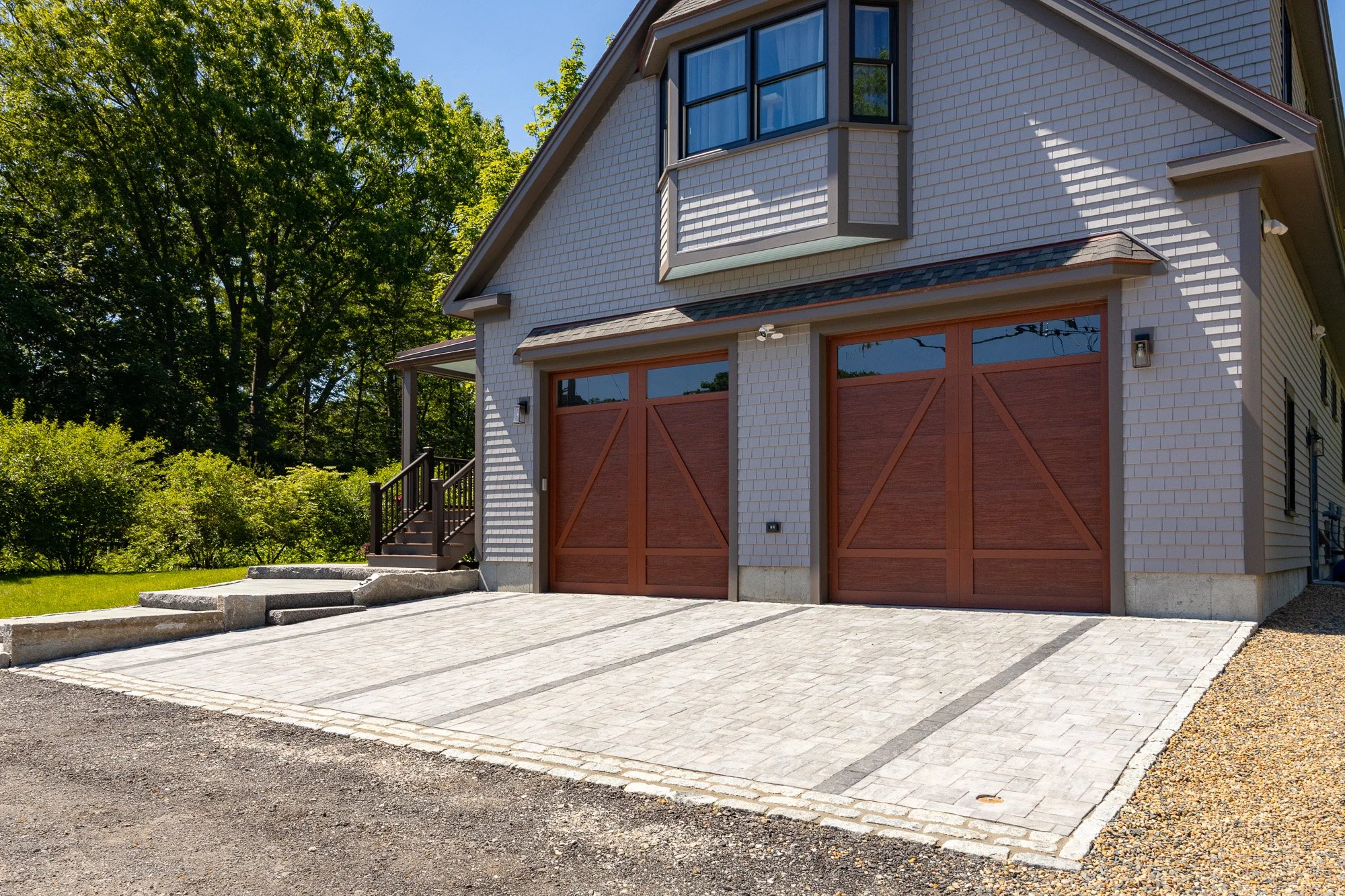 Front view of a house with white siding, large garage with wooden doors, and a paved driveway. There are stairs leading to a side entrance, trees in the background, and a bright, sunny day.