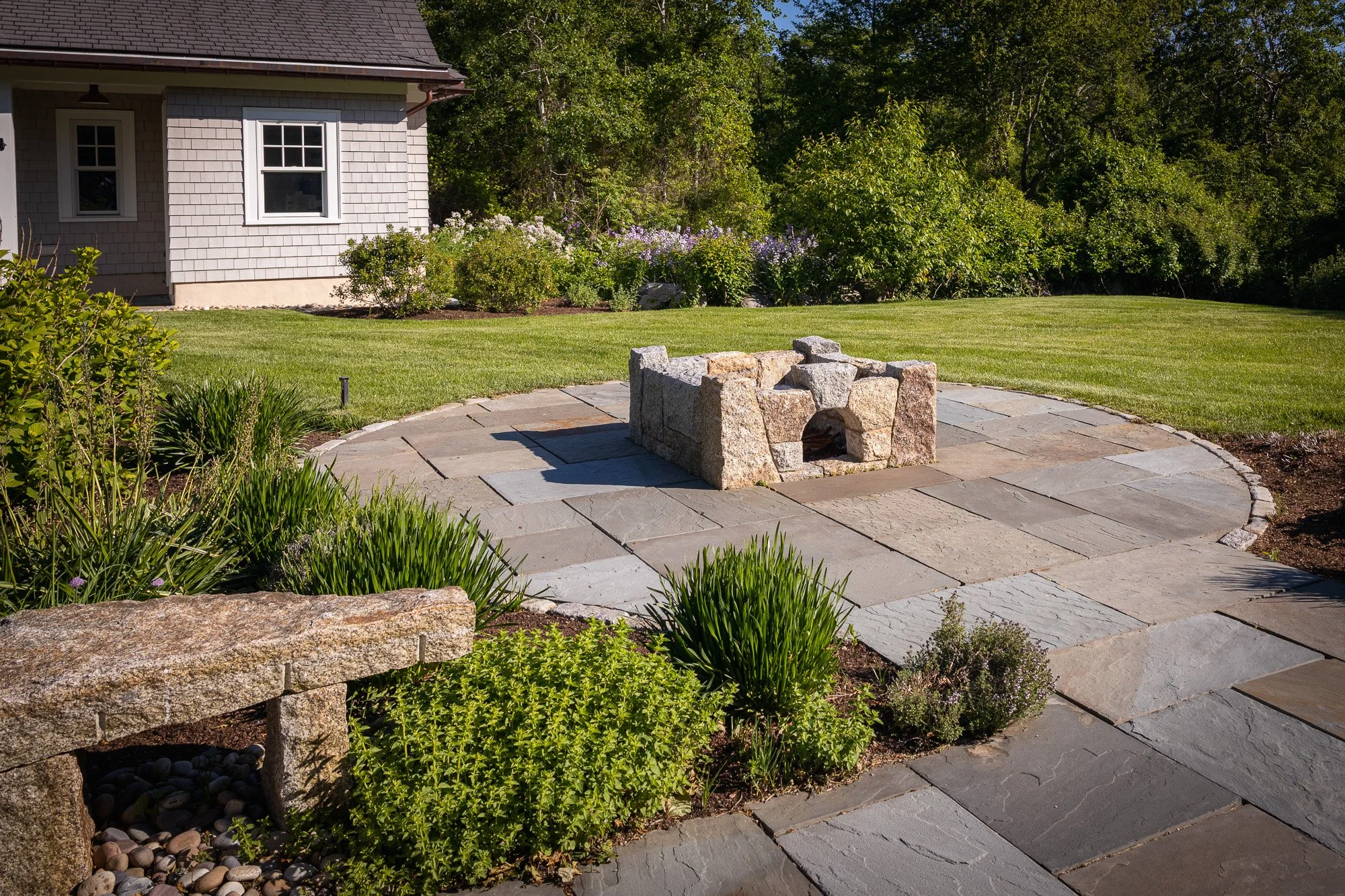 A backyard with a stone fire pit on a paved circular patio, surrounded by green grass, bushes, and flowering plants, with a house partially visible on the left and trees in the background.