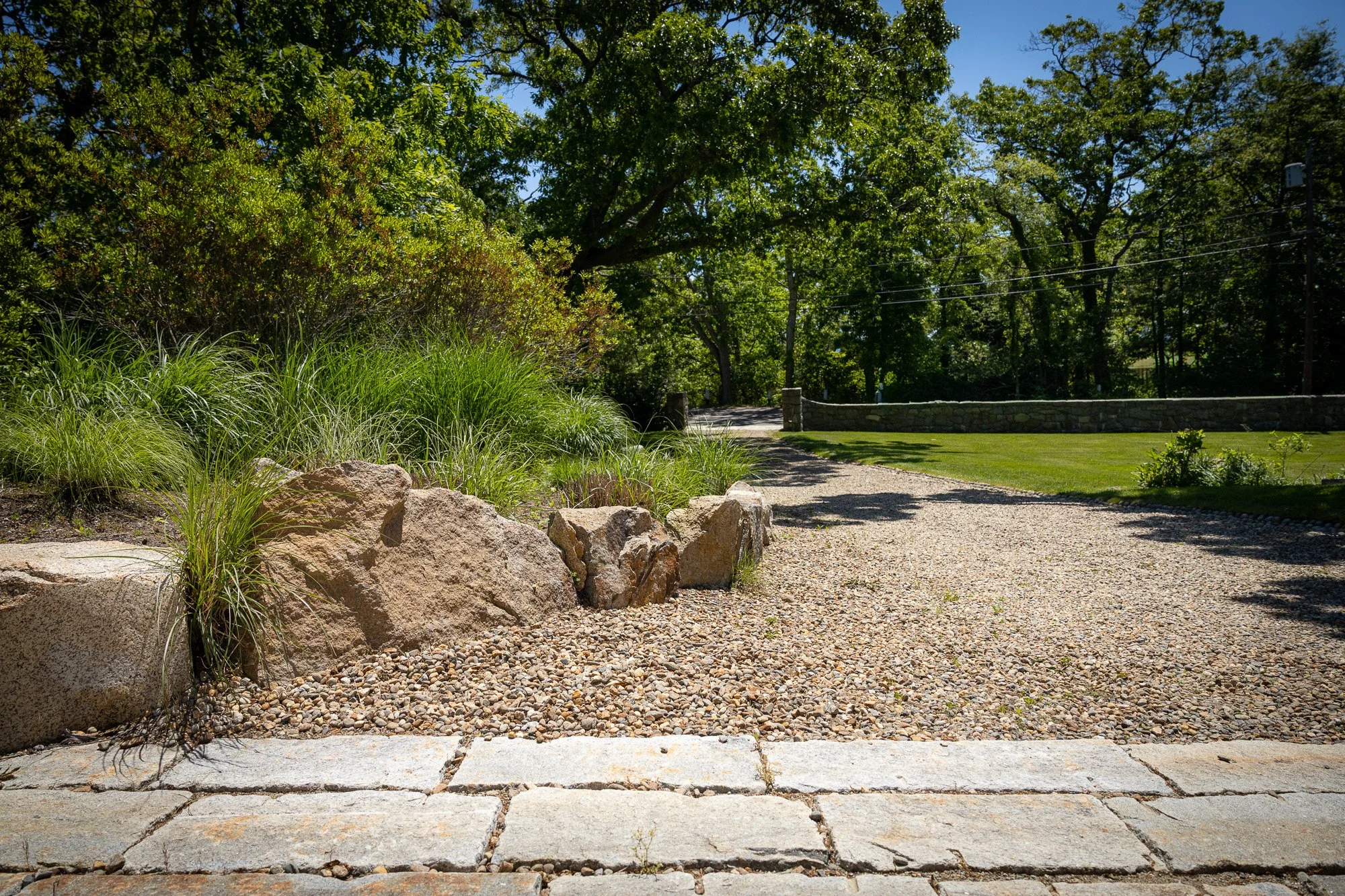 A garden scene with large rocks, green grass, bushes, tall trees, and a stone wall with a pathway near it on a sunny day with a clear blue sky.