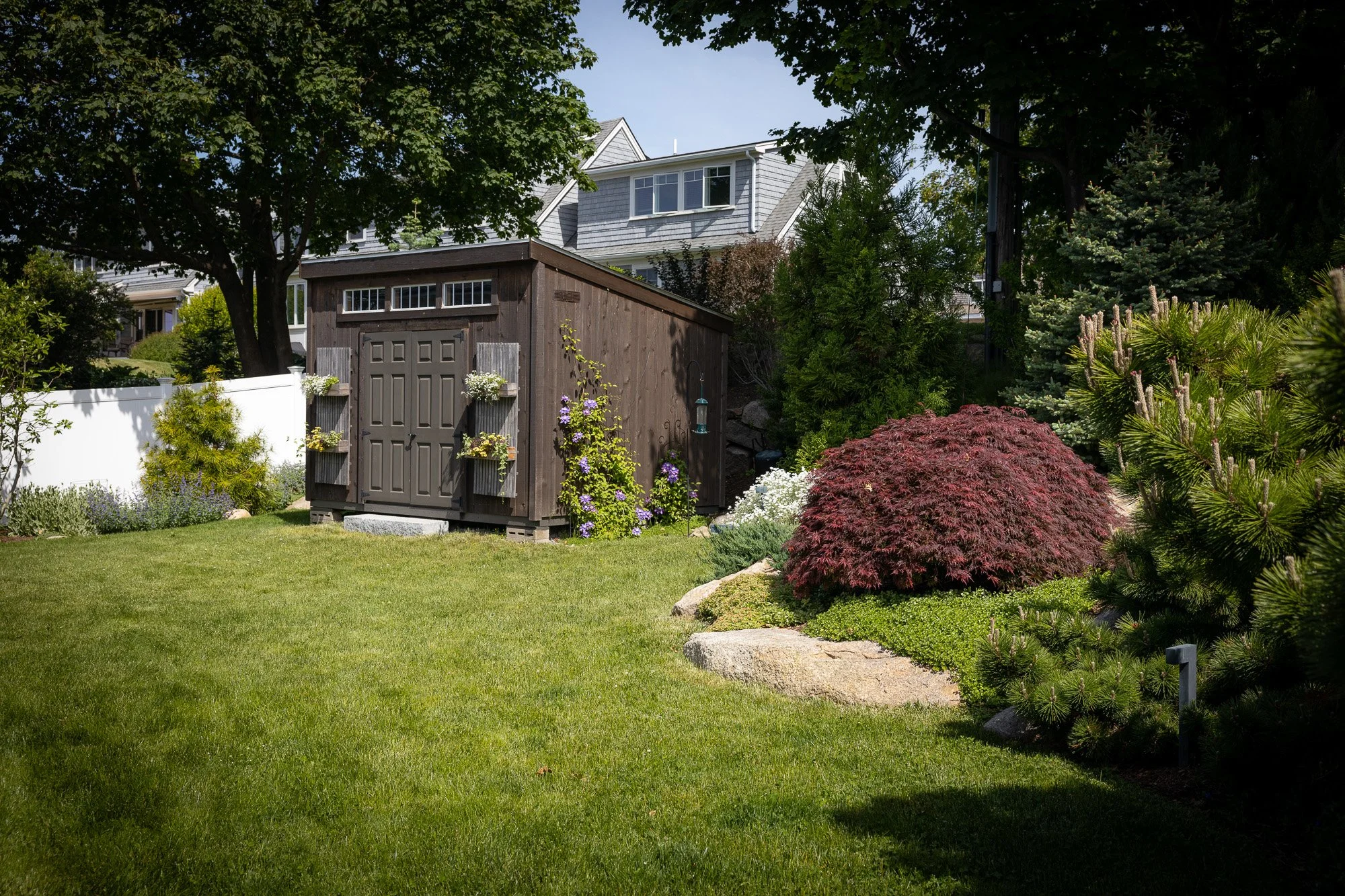 A lush backyard garden with green grass, colorful bushes, and tall trees in front of a gray house with a sloped roof.