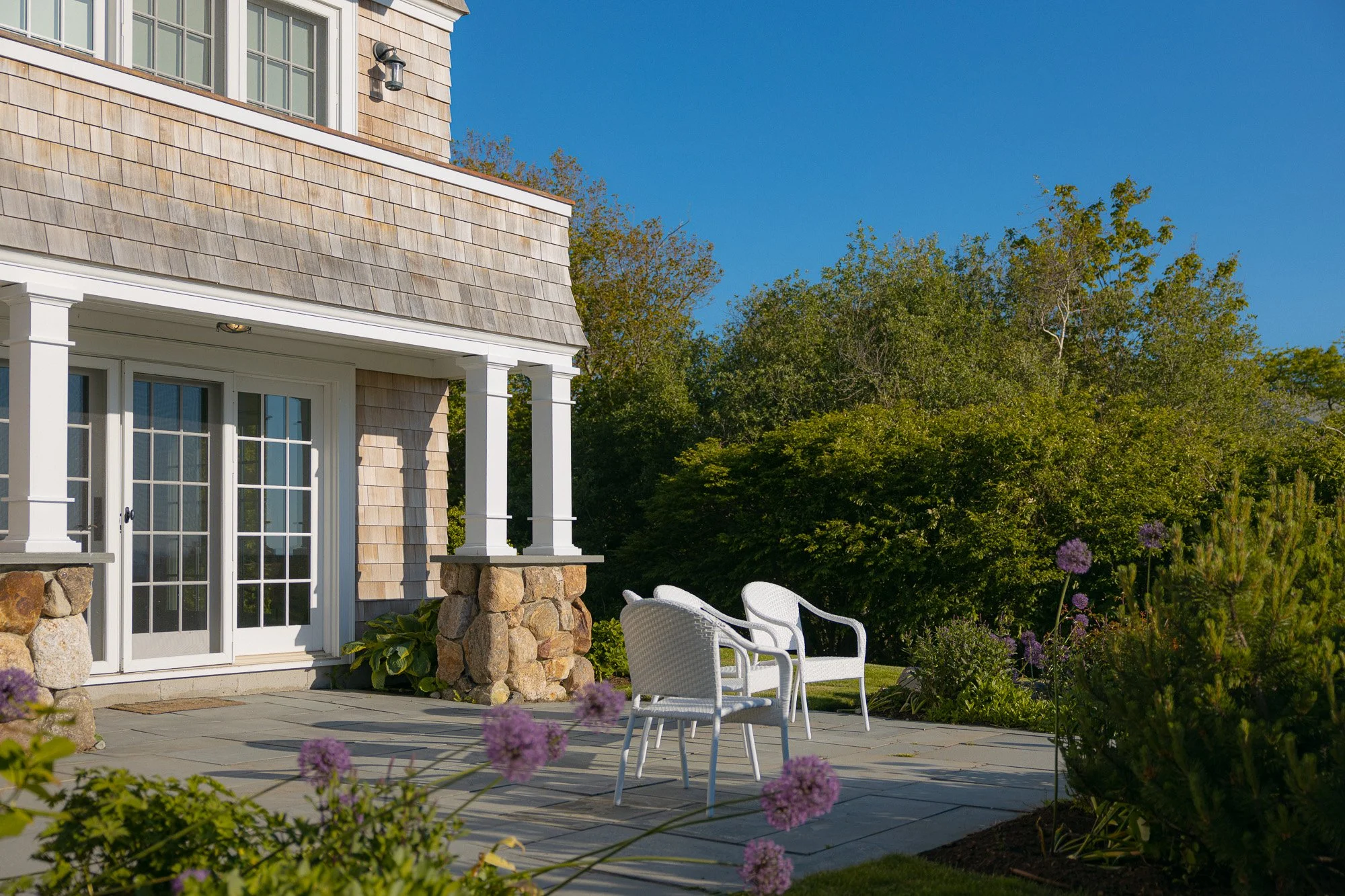 A backyard patio area with white outdoor chairs, a house with beige shingle siding, glass sliding doors, and a porch with stone pillars. There are green trees and flowering plants around, and the sky is clear and blue.