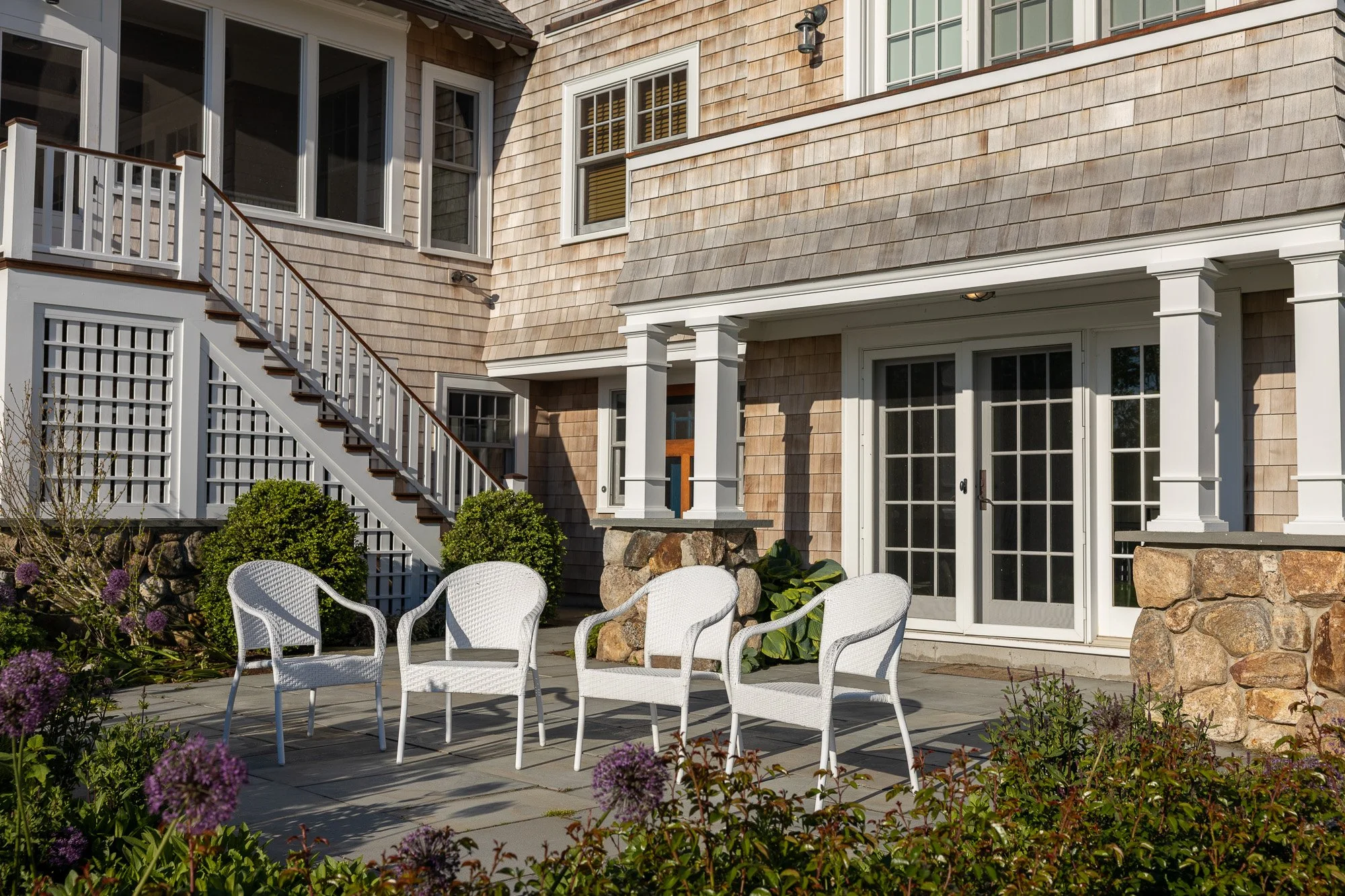 Backyard patio with white chairs, surrounded by purple flowers and green bushes, attached to a house with wooden shingles, large windows, glass doors, and a staircase leading up to a balcony.