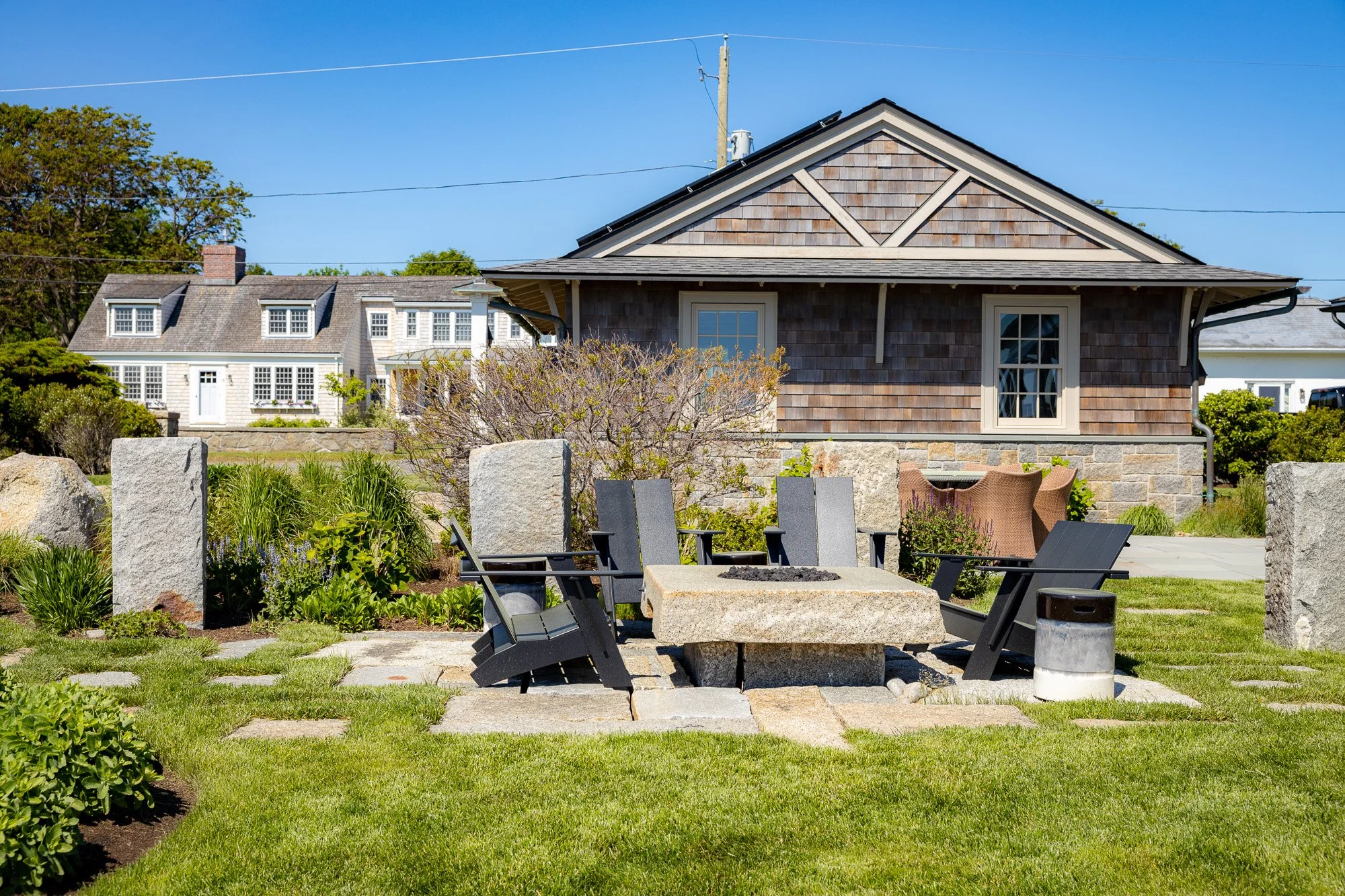 Outdoor patio with black chairs surrounding a stone fire pit, landscaped garden with bushes and trees, and houses in the background under a blue sky.