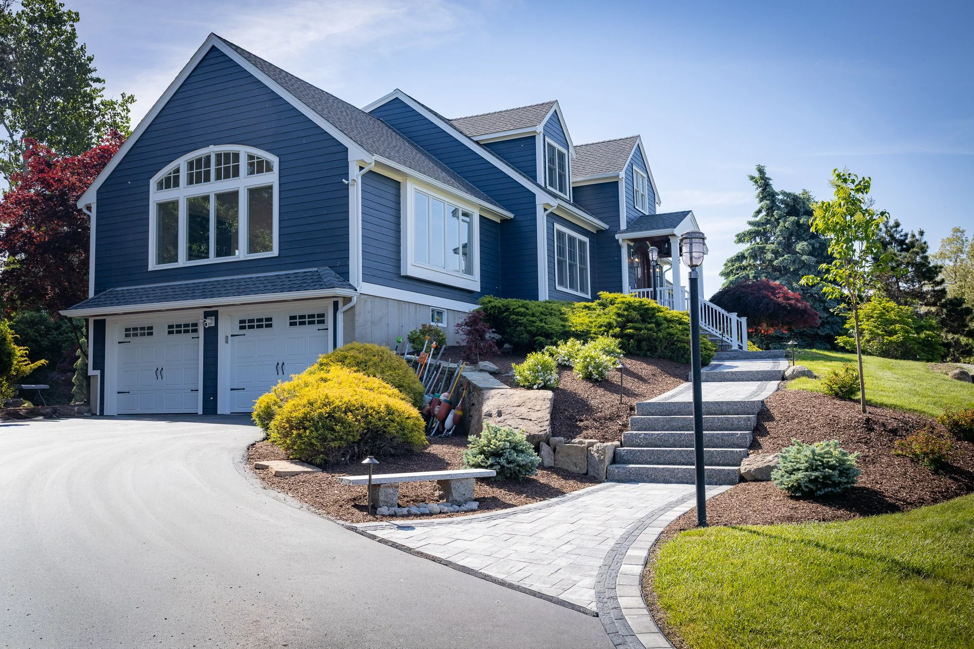 A two-story house with blue siding, white trim, and a gray shingle roof. There is a garage at the front with a white door, and a short walkway with steps and landscaping leading to the front porch, surrounded by trees, bushes, and a manicured lawn.