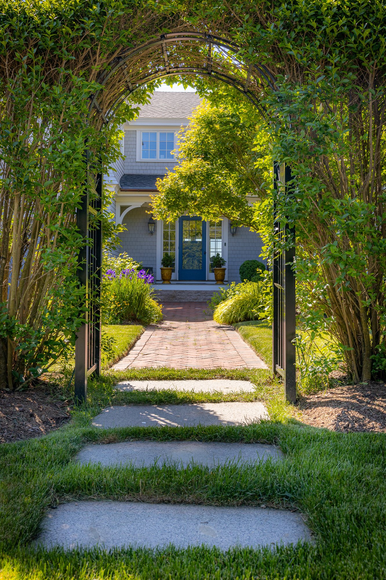 View of a house with gray siding, white trim, and a front porch, seen through a black metal archway framed by green bushes and trees, with a brick walkway leading to the front door.