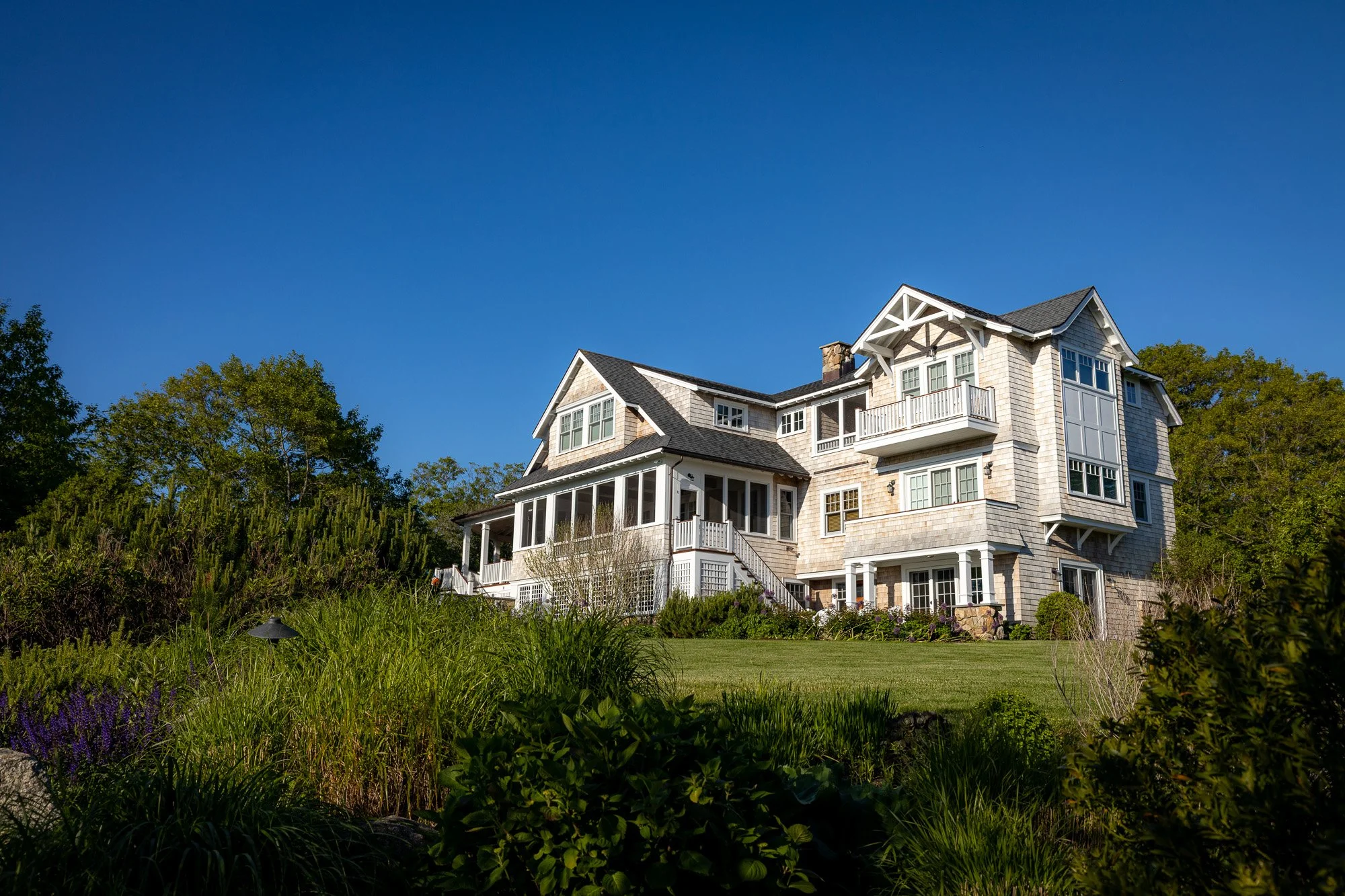 A large, multi-story house with a gabled roof, large windows, and a balcony, surrounded by greenery and trees under a clear, blue sky.