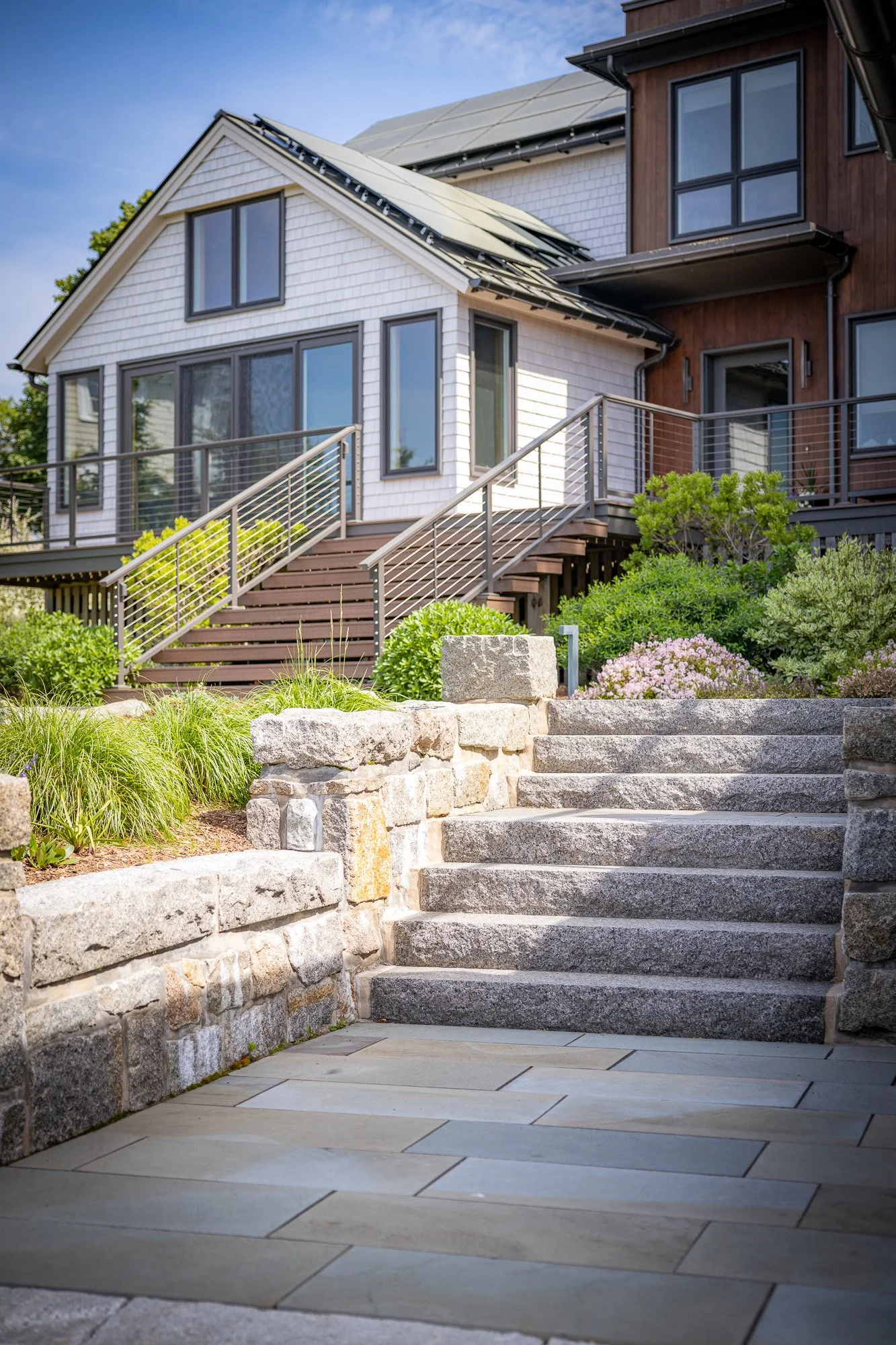 Exterior view of a modern multi-story house with a white and brown facade, large windows, and a staircase leading to the entrance. The house has solar panels on the roof, landscaping with bushes and flowers, and a stone retaining wall at the bottom o