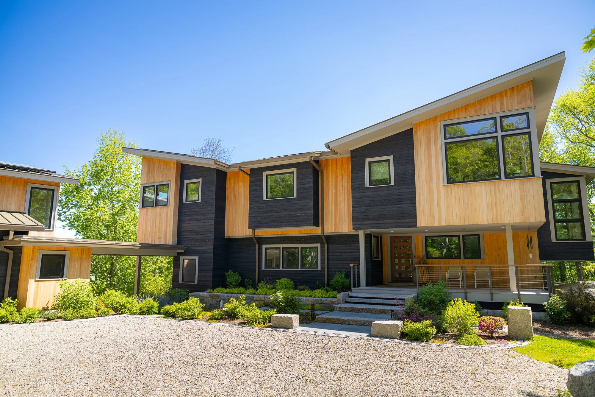Modern multi-story house with large windows, wooden and black exterior, surrounded by greenery and a gravel pathway.