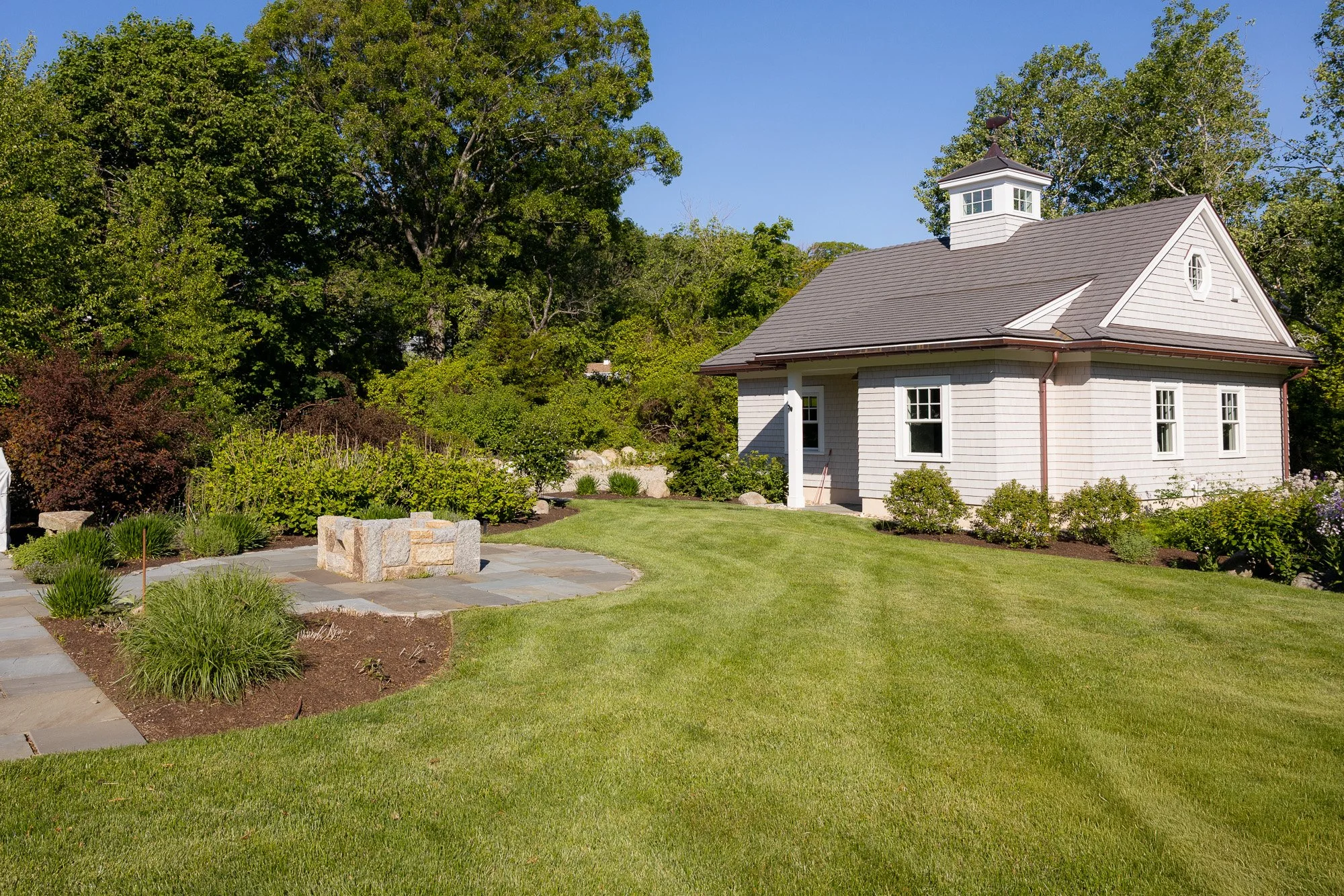 A white house with a gray roof and small white windowed tower on top, surrounded by green trees and well-maintained lawn, with a stone patio area in the foreground.
