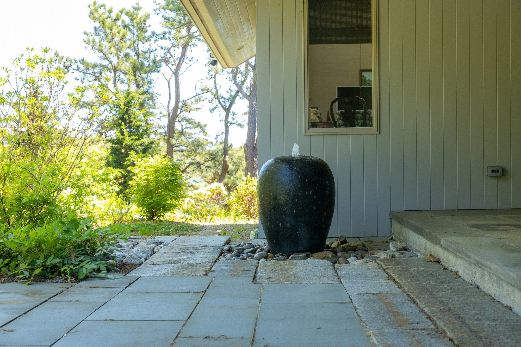Exterior view of a house porch with a black water fountain, stone path, greenery, and trees in the background.