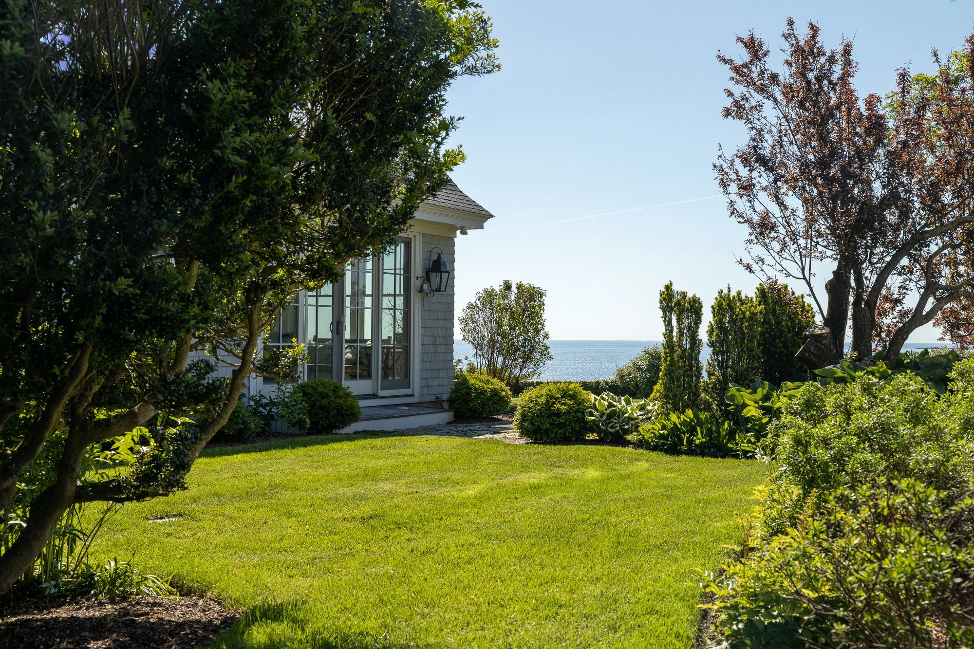 A backyard with a green lawn, surrounded by bushes and trees, and a house with large glass doors and an outdoor light fixture, overlooking the ocean on a clear, sunny day.