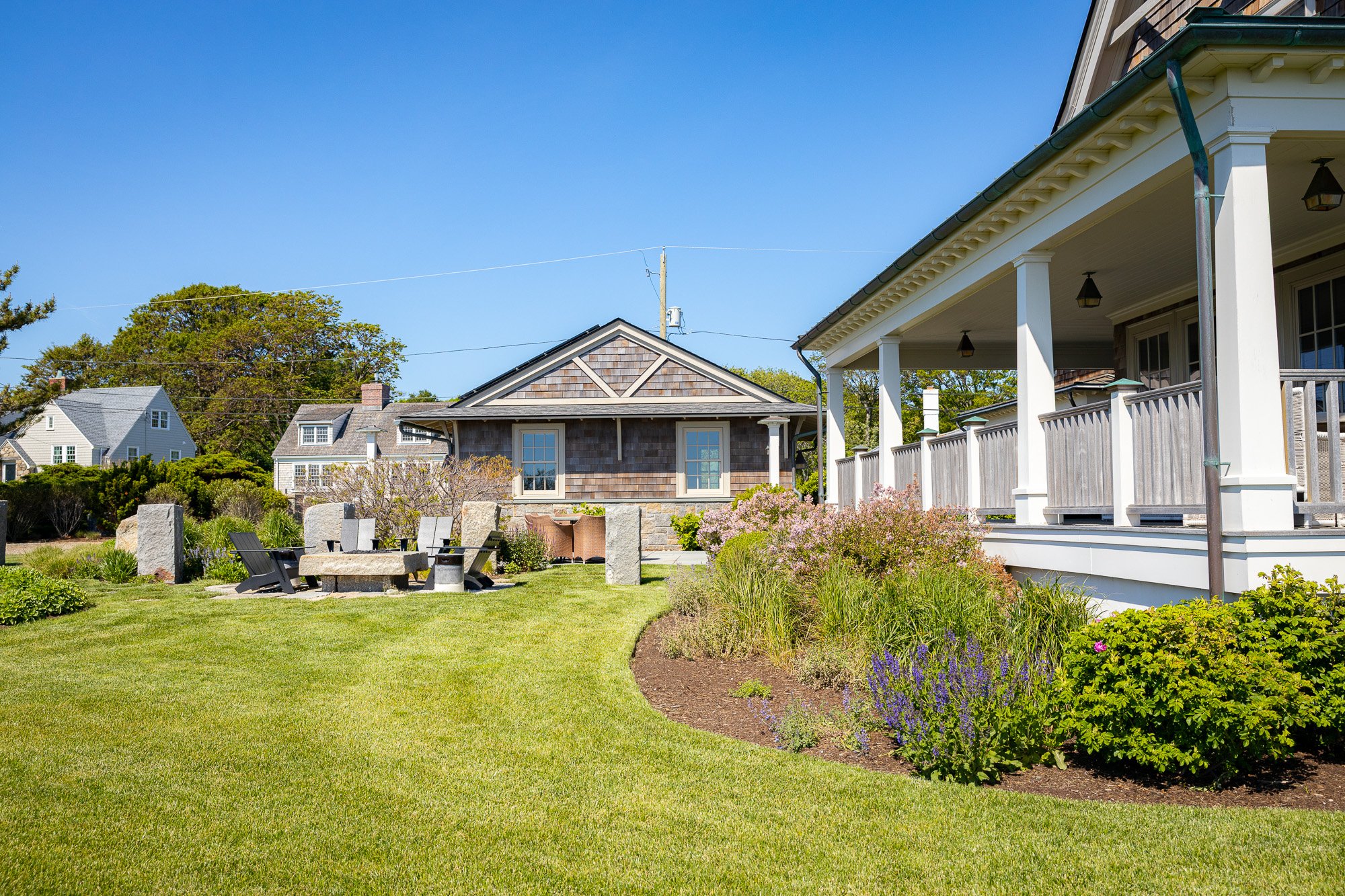 A backyard garden with a green lawn, flower beds, stone pathways, and outdoor seating, surrounded by houses with porch areas, trees, and a clear blue sky.