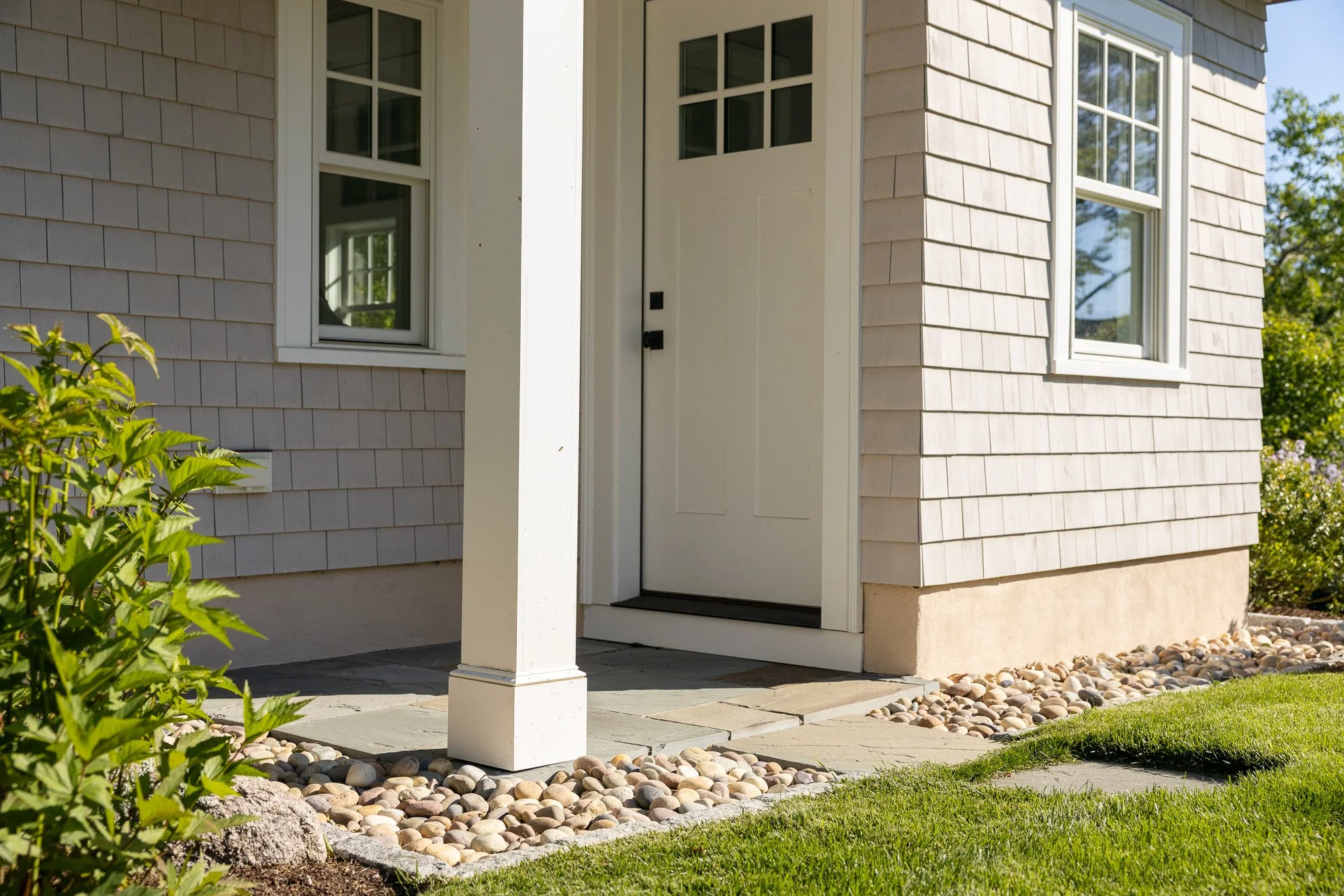 The front porch of a house with two windows, a white door, and a supporting column. The house is clad in light beige horizontal siding, and the porch has a concrete slab with a border of smooth rounded stones. Green bushes and grass surround the porc