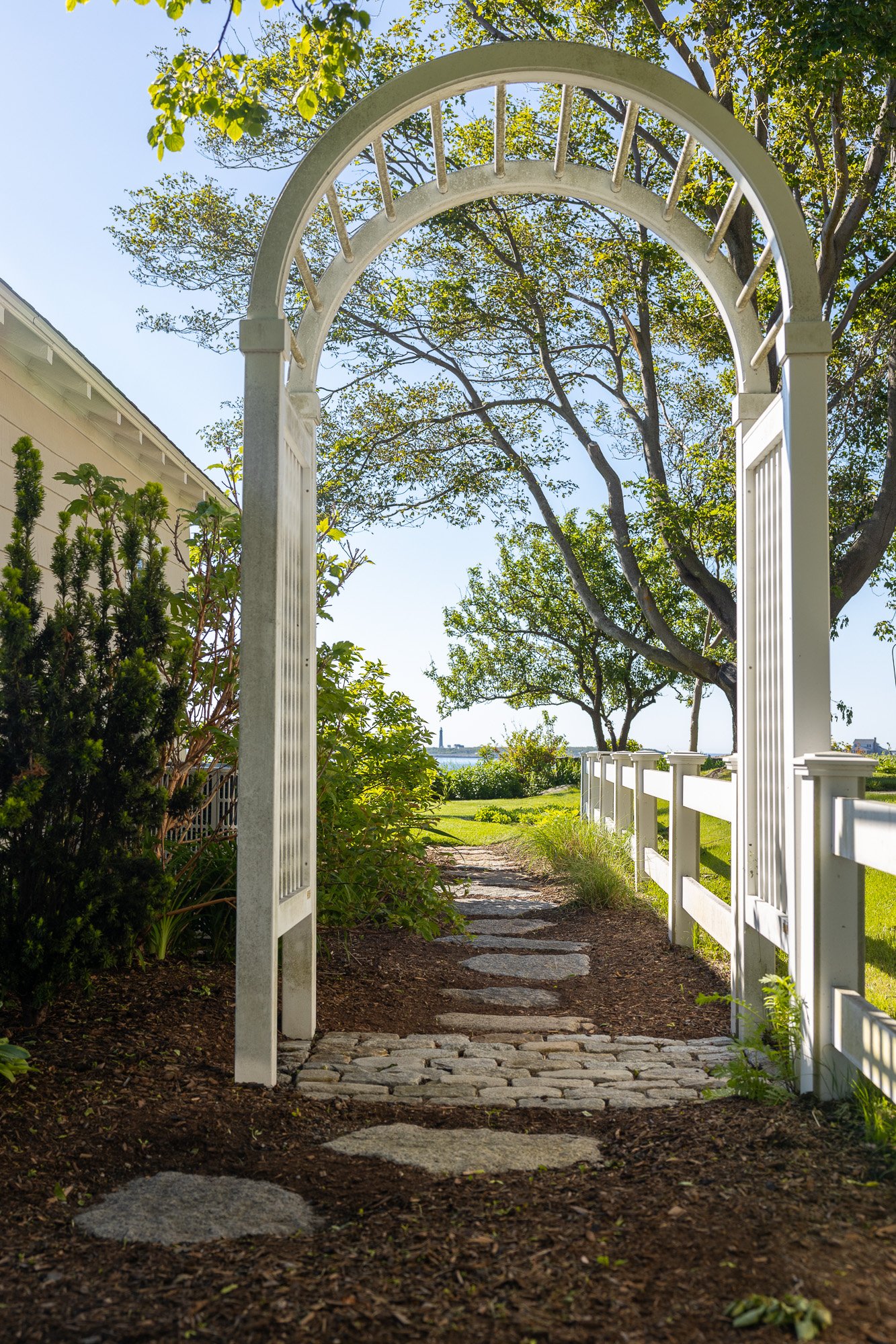 A white garden archway with decorative detail, framing a stone pathway leading through a green garden to a body of water in the distance. There are trees and a white fence along the path under a clear blue sky.