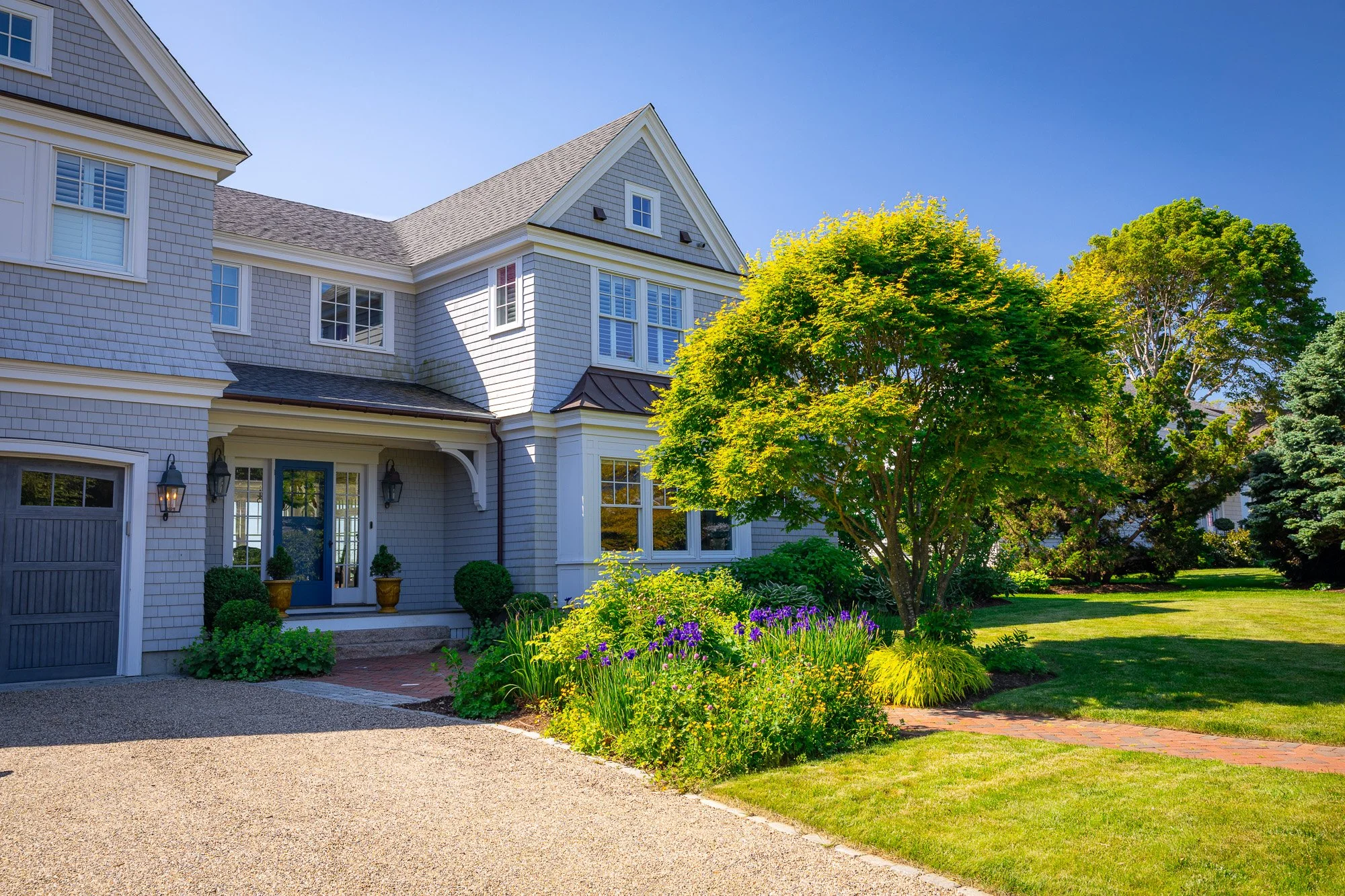A large, light gray house with a peaked roof, multiple windows with white shutters, and a front door with outdoor lanterns. The front yard features a well-maintained lawn, a tree with lush green foliage, and a flower bed with purple and yellow flower