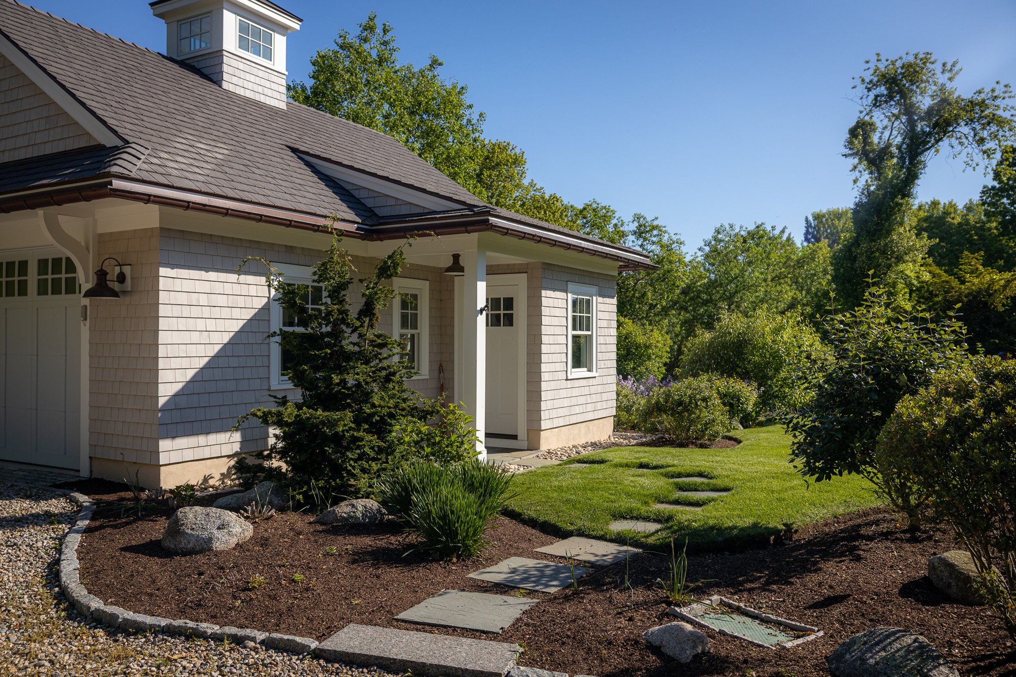 A house with a shingle roof, white window frames, and a white door, surrounded by a well-maintained garden with various plants, bushes, trees, and a stone-paved pathway under a clear blue sky.