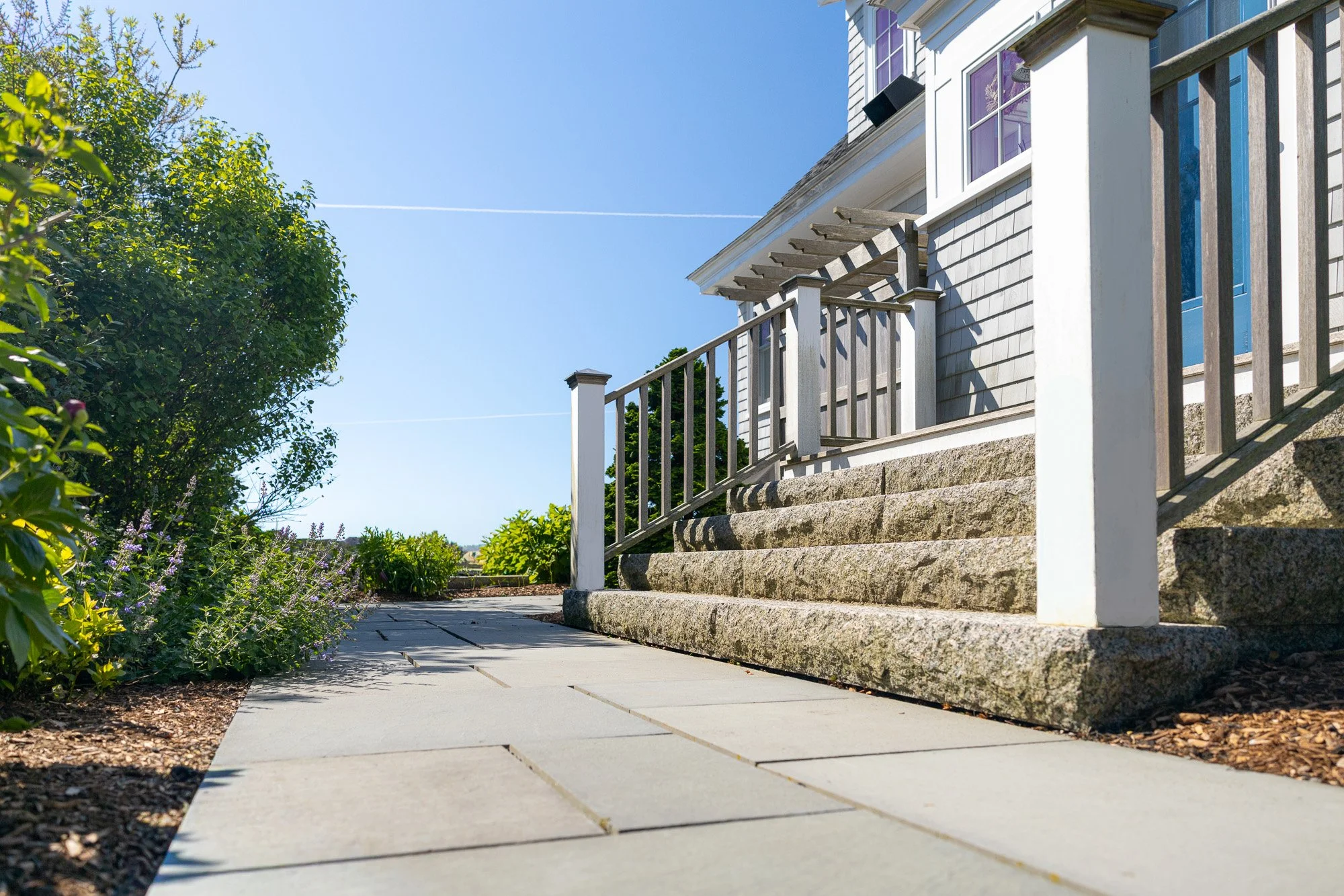 Side view of a house with a staircase leading to a front door, pathway, garden with plants and flowers, and blue sky with contrails.