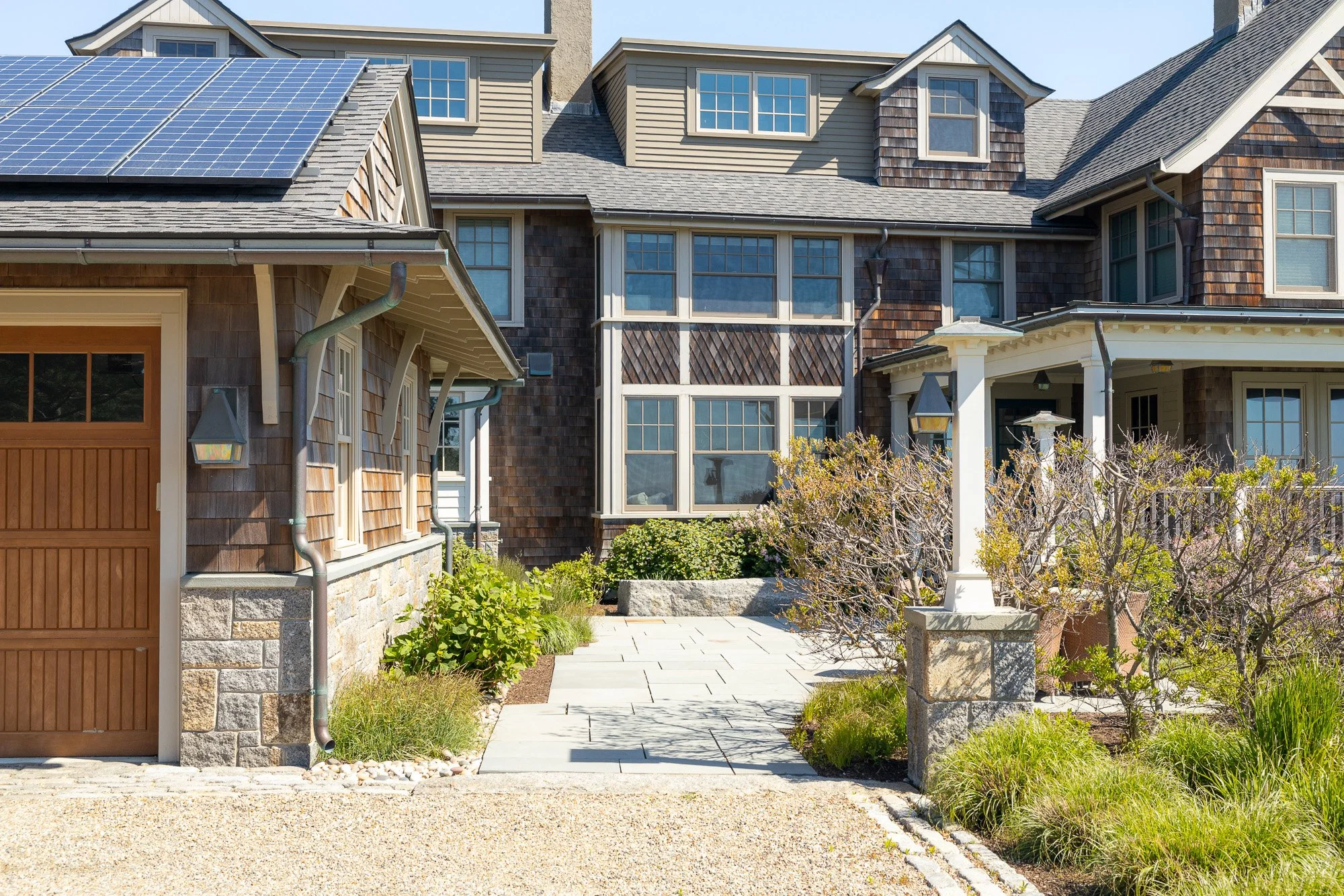 The image shows the front of a large, multi-story house with brown wooden siding and shingles, shed dormer windows, and a roof equipped with solar panels. There is a stone pathway leading to the house entrance, flanked by landscaped bushes and plants