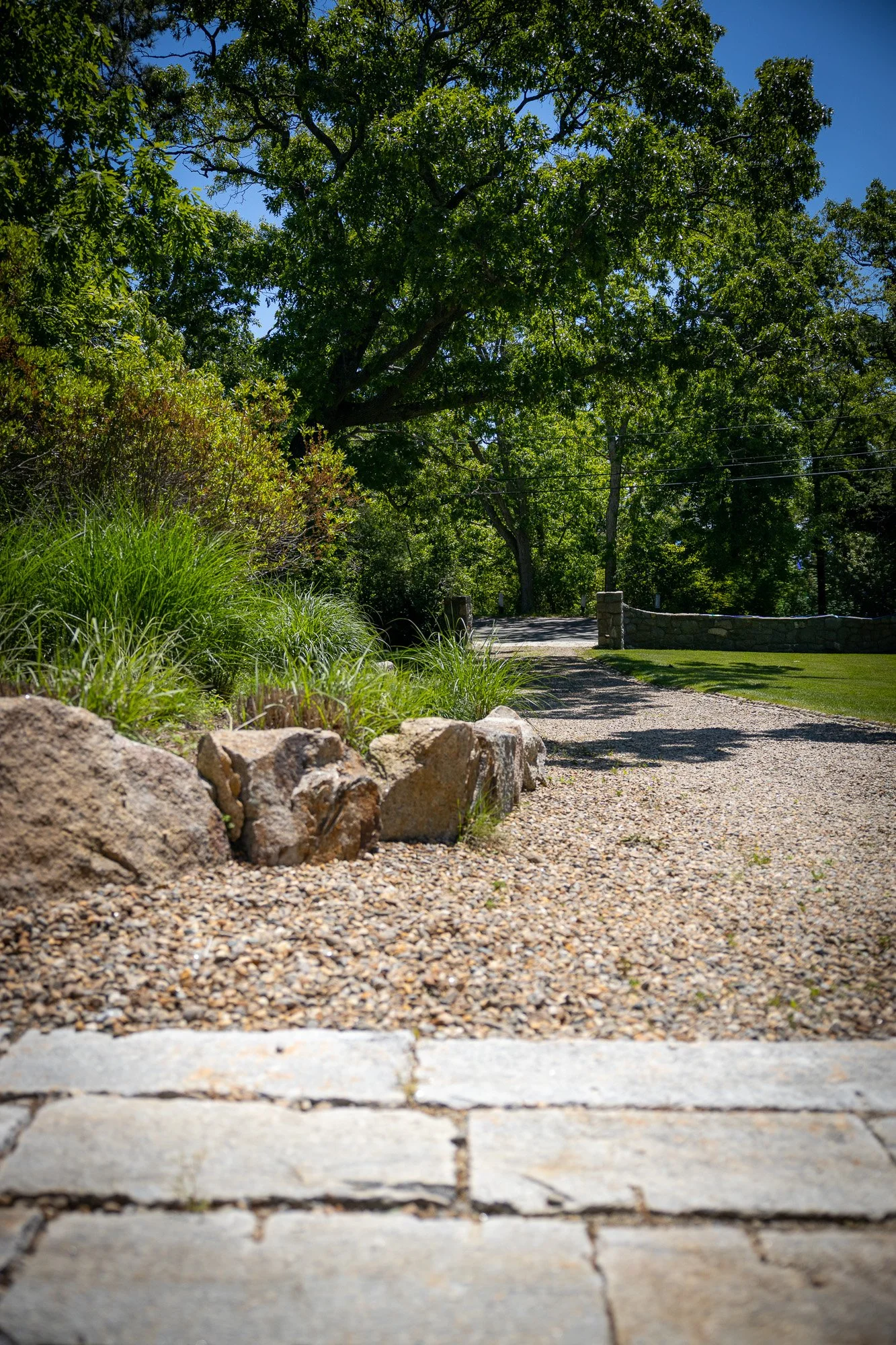 View of a gravel pathway with grass and large rocks on the left side, trees in the background under a blue sky.