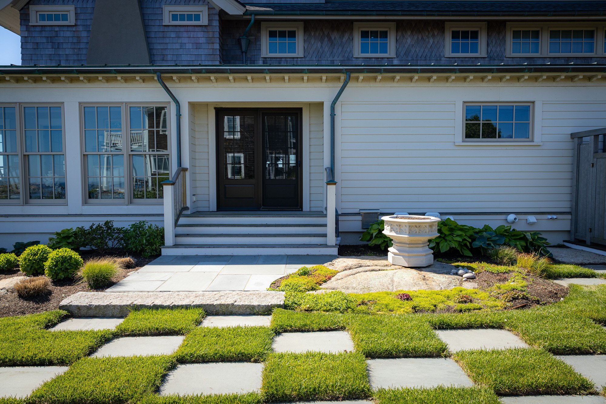 Front yard of a house with a set of stairs leading to a black door, surrounded by a well-maintained garden with green bushes, a stone pathway, and decorative elements including a white planter with plants.