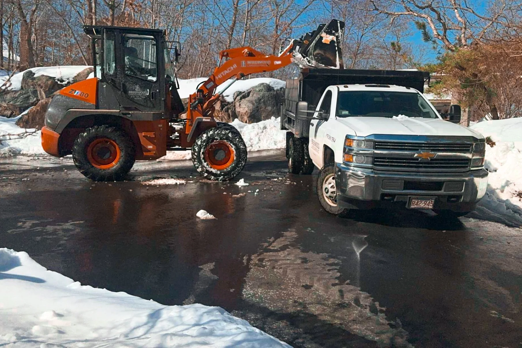 A snow-covered parking lot with a large orange front loader lifting a snowplow attachment toward the bed of a white pickup truck. Snow and ice are visible on the ground and surrounding trees, with a clear blue sky overhead.