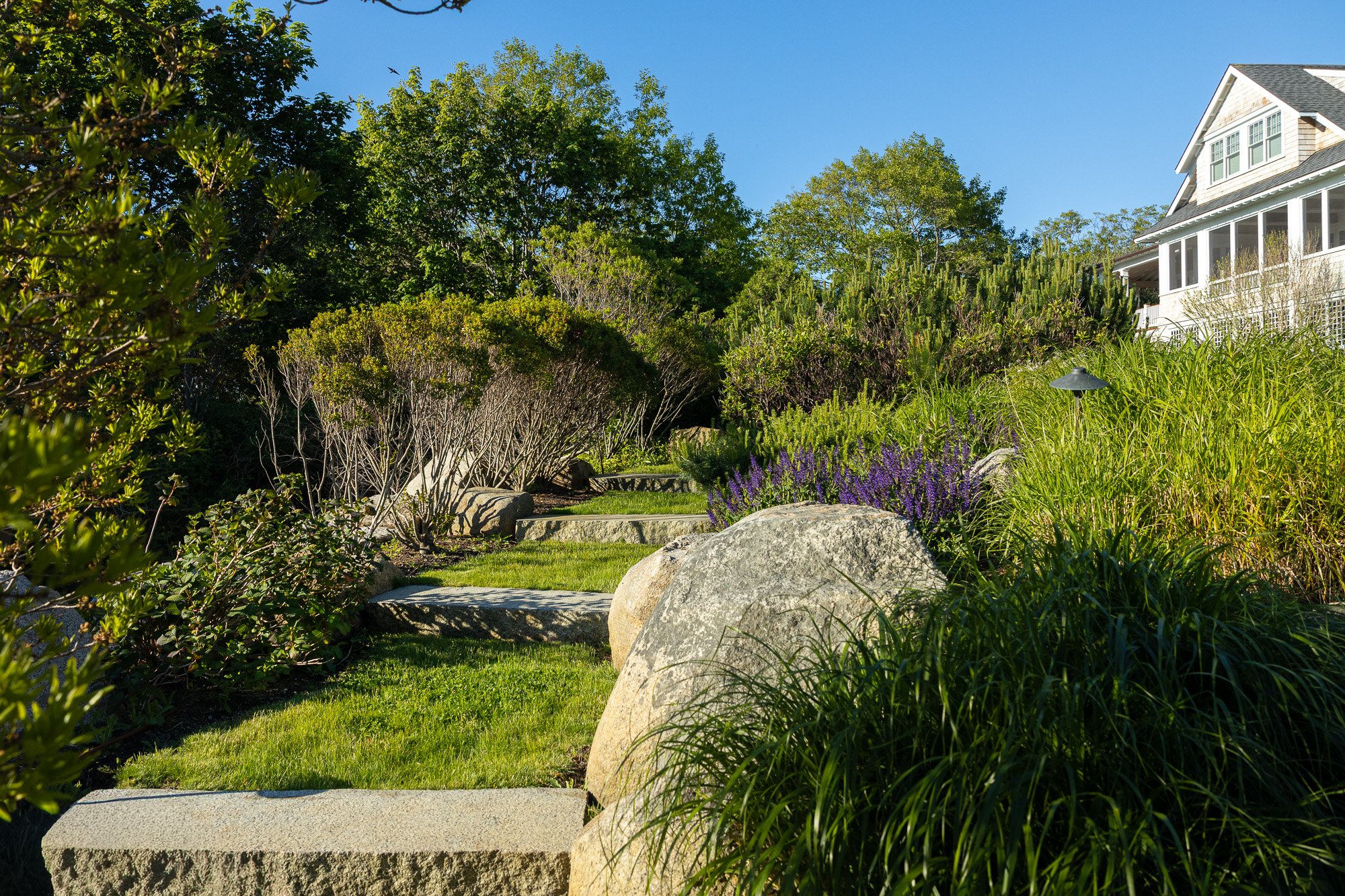 A lush, landscaped garden with large rocks in the foreground, a manicured lawn, dense bushes, and tall trees under a clear blue sky, with a white house partially visible on the right.