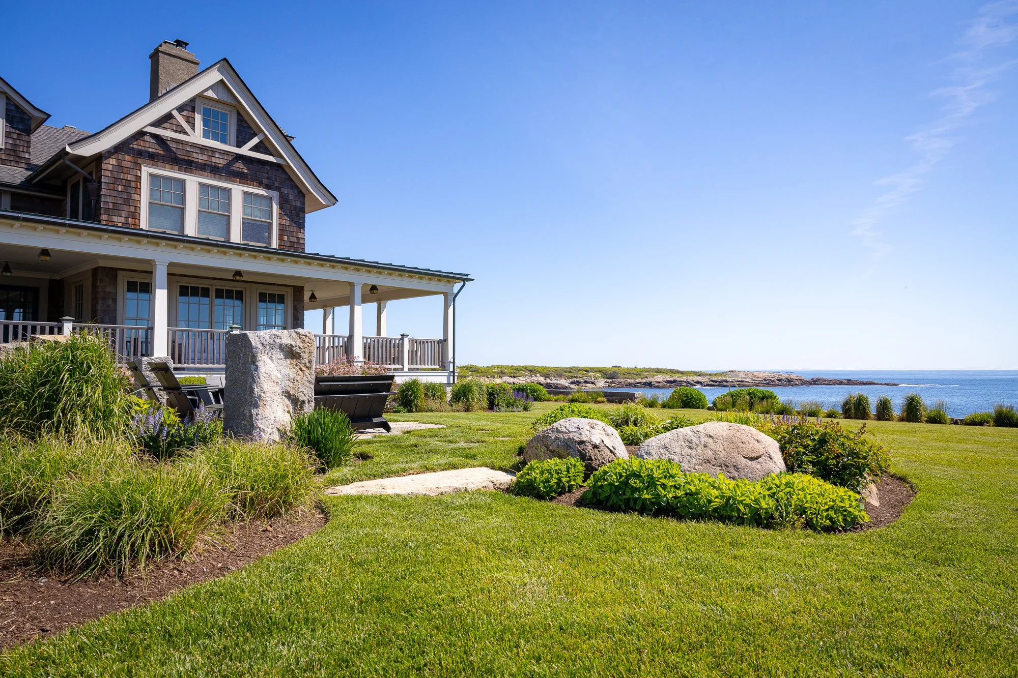A large house with a wooden exterior and multiple windows, surrounded by a well-maintained garden with rocks and green plants, overlooking a coastal landscape with rocks and water.