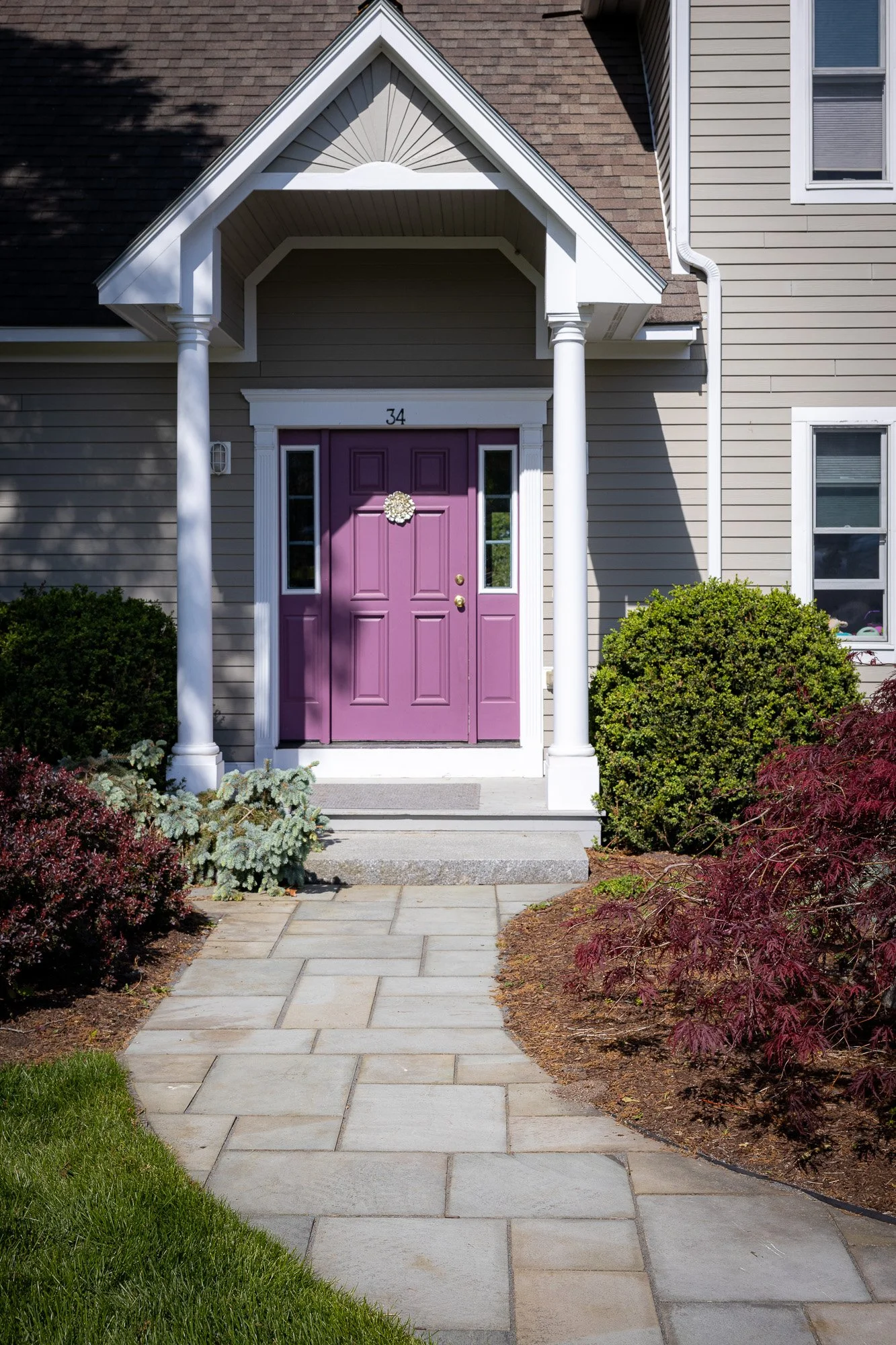 Front view of a house with a purple front door, two white columns supporting a small roof above the door, and surrounded by greenery and bushes.