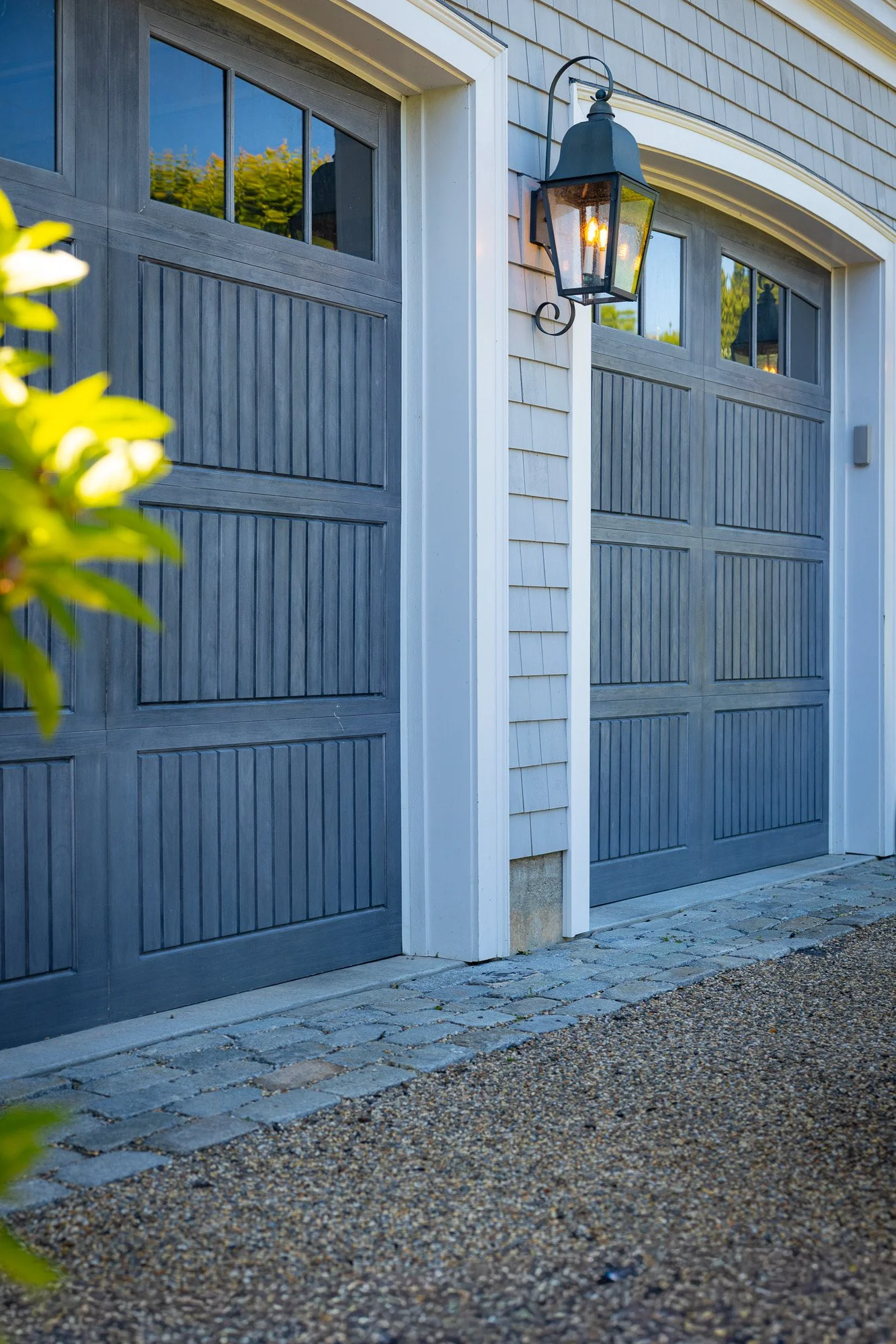 A close-up of a gray garage door with decorative window panels at the top, mounted on a house with beige siding and white trim. There is a black outdoor lantern light fixture turned on, illuminating the area beside the garage. The ground is composed 