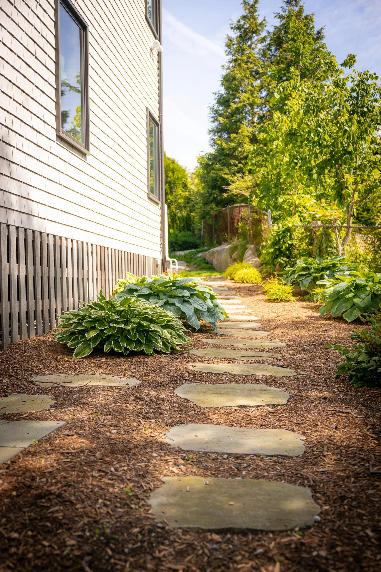 Stone pathway leading through a garden with green plants on either side, next to a house with beige siding and white trim. The background includes trees and a bright blue sky.