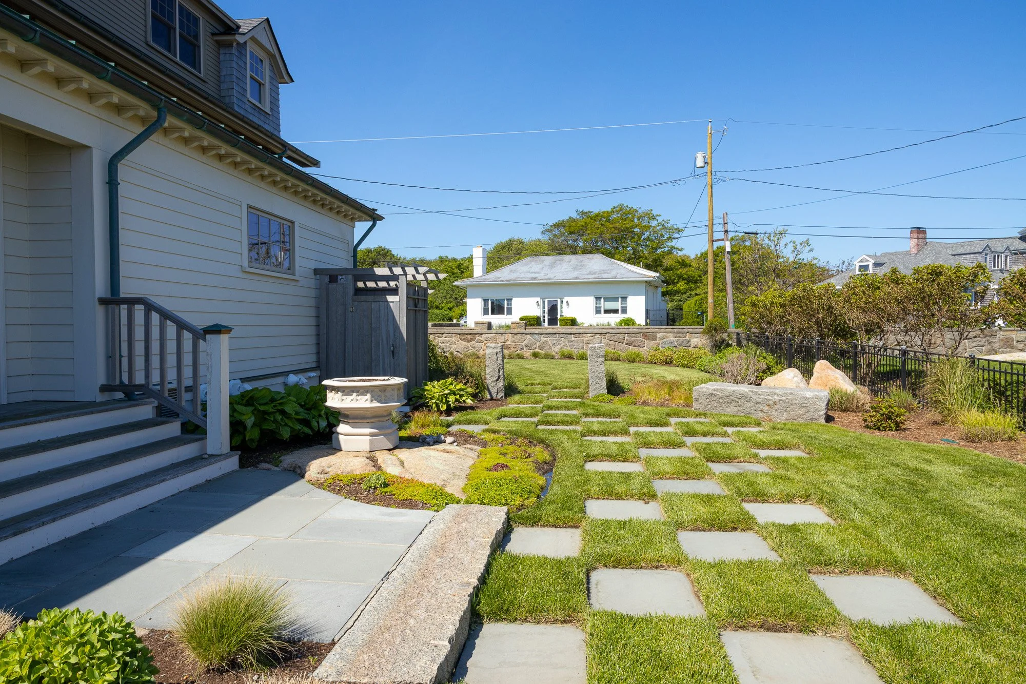 View of a backyard garden with grass and stone pathway, neighboring houses in the background, and a partly cloudy blue sky.