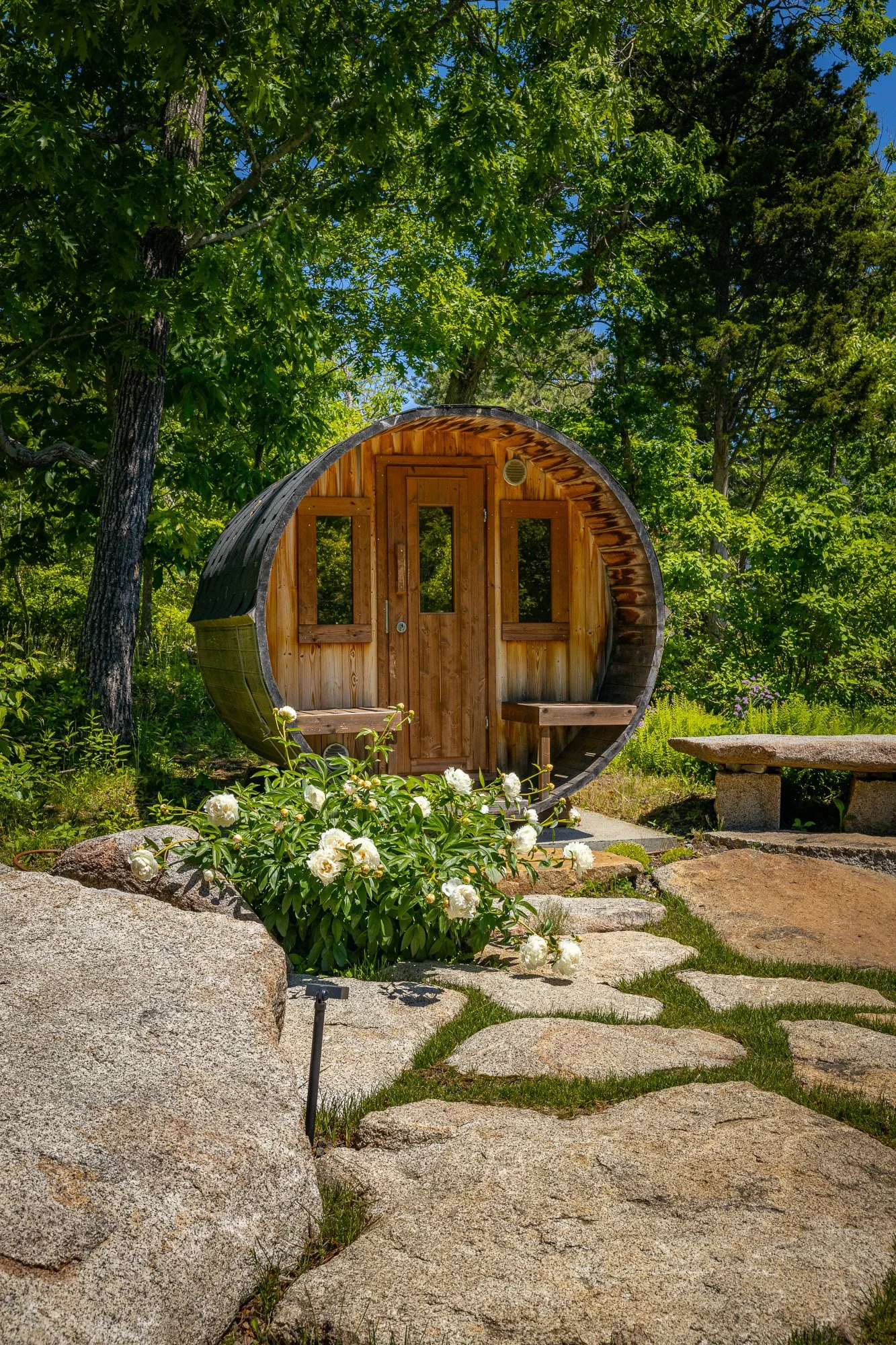 A small wooden house with a semi-circular roof, surrounded by greenery, rocks, and flowers in a garden setting.