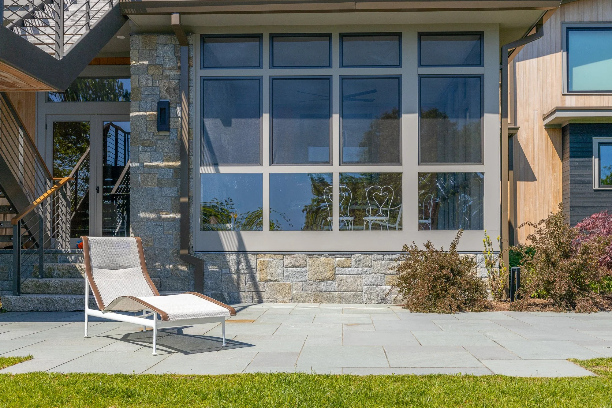 Backyard patio with a white lounge chair, a glass table, and a house with large windows, stone, and wood exterior.