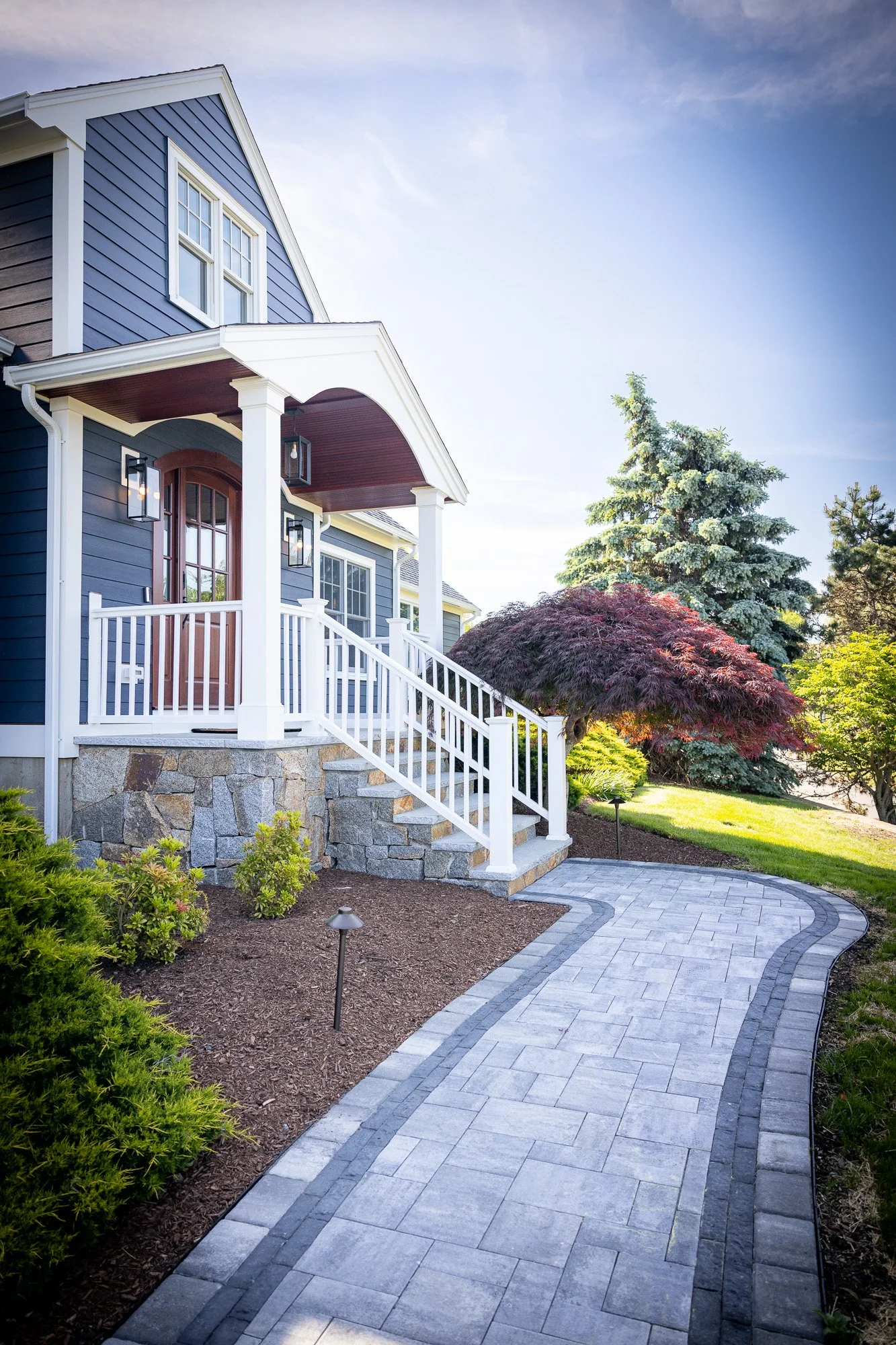 A blue two-story house with white trim, a covered front porch, and a pathway leading to the yard. The yard has various bushes, a red-leafed tree, and a large evergreen tree under a clear blue sky.