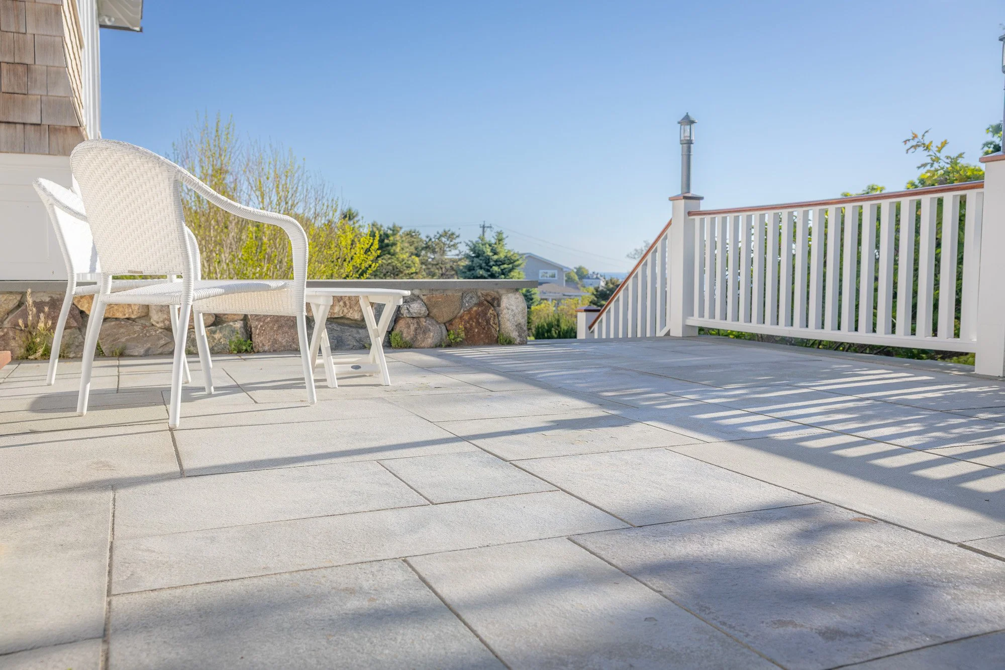 A spacious outdoor patio with white wicker chairs and a table, overlooking a scenic neighborhood with trees and rooftops, under a clear blue sky.