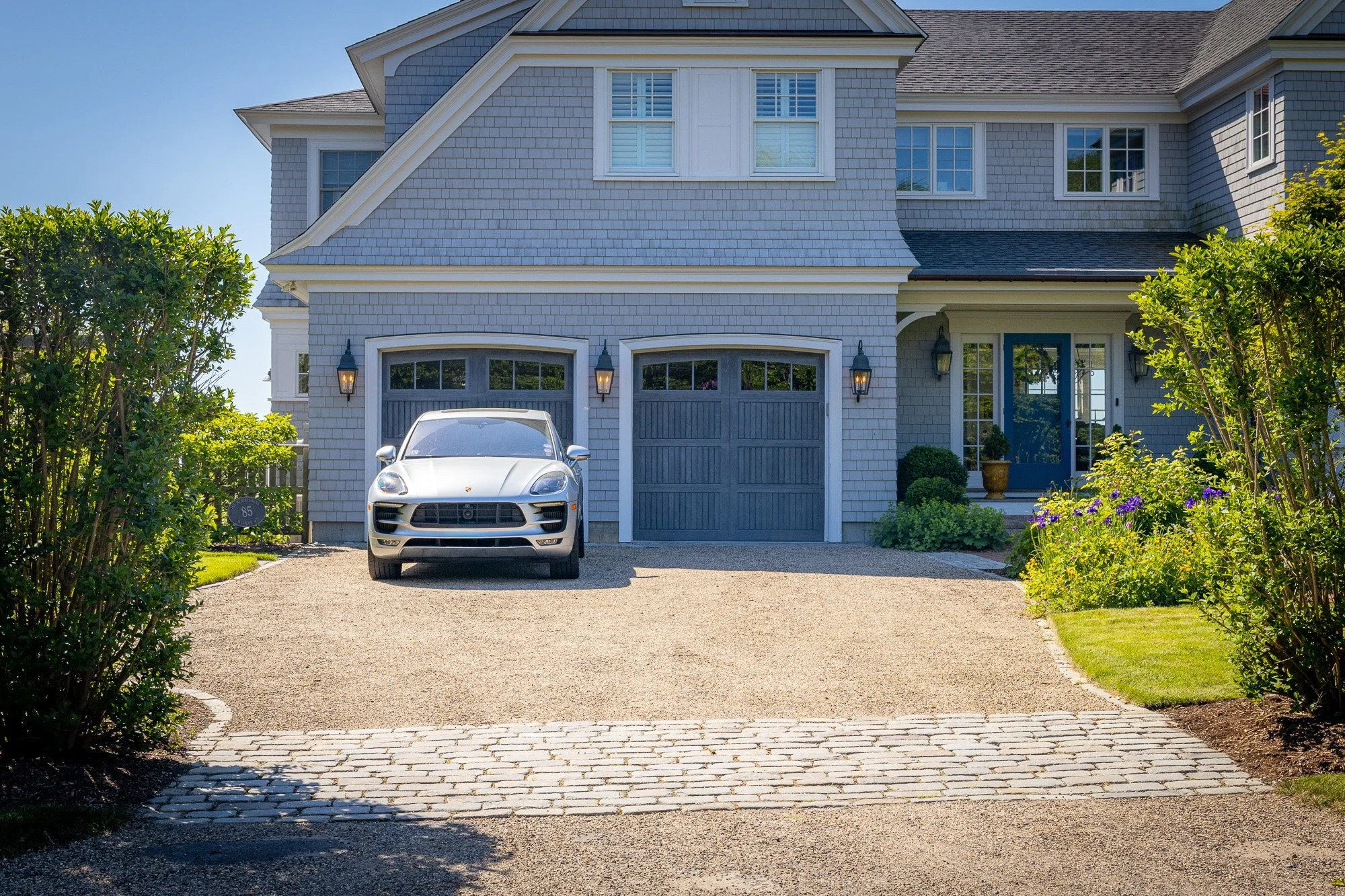 Front view of a light blue, two-story house with a garage, white trim, and a driveway with a silver car parked, surrounded by green bushes and colorful flowers.