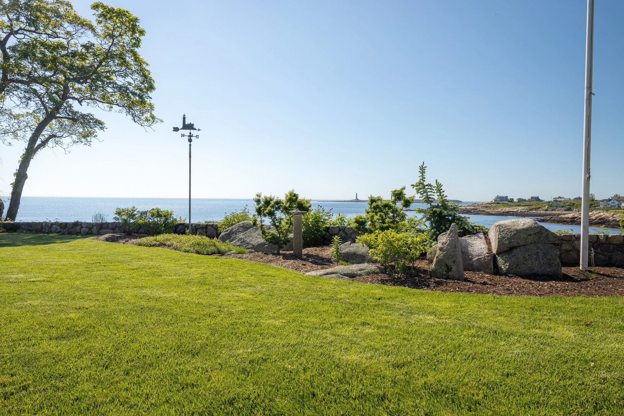 A landscaped seaside yard with a green lawn, trees, a stone border, and a view of water and a lighthouse in the distance.
