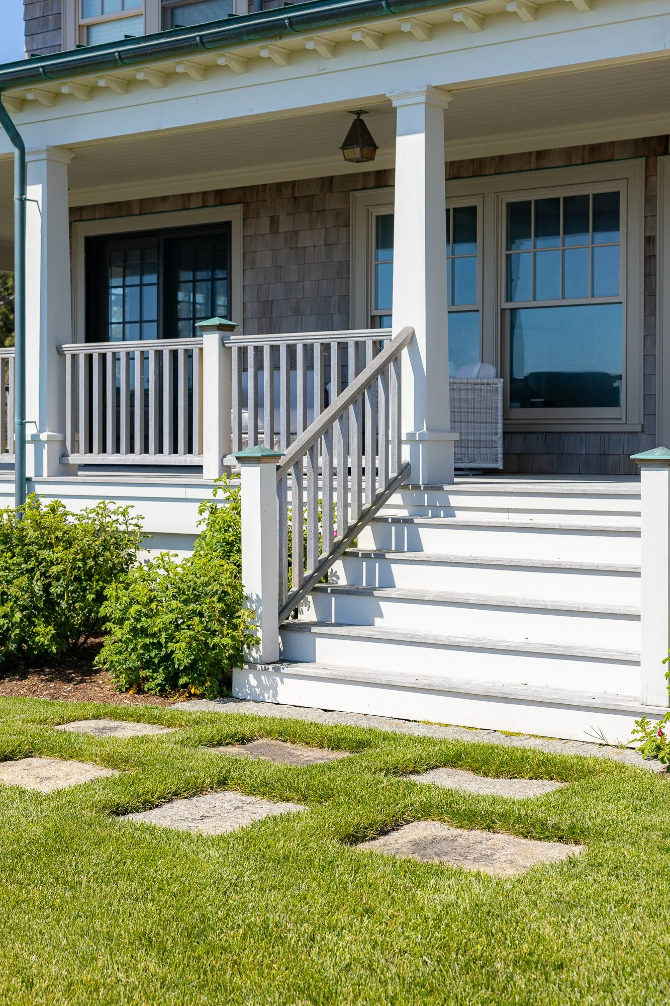 Front porch of a house with white steps, railing, and posts, leading to a sliding glass door, and a large window with a wicker chair nearby. The house has a green roof and a small overhang supported by columns. In front of the house, green grass with