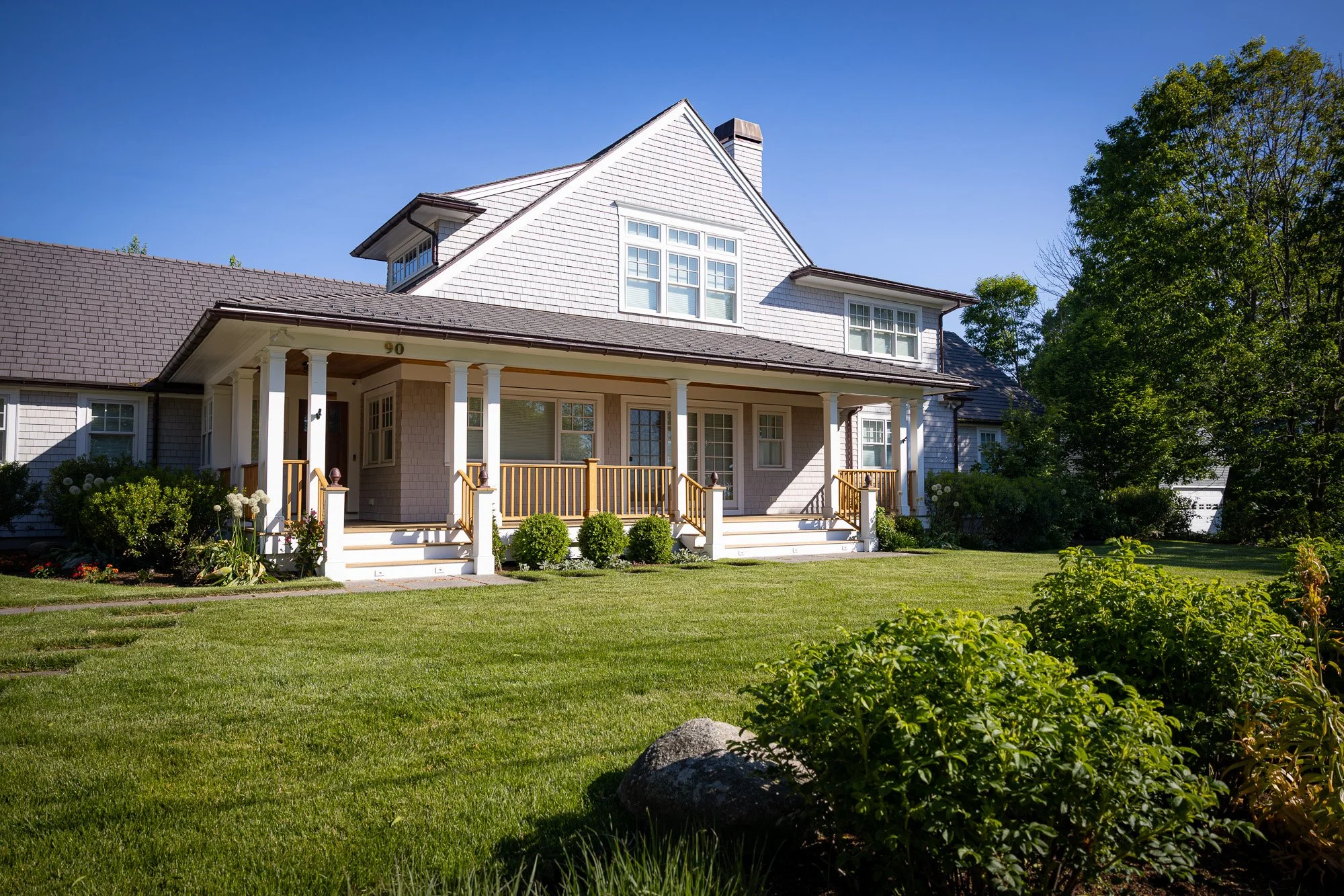 Front view of a two-story house with light-colored siding, multiple windows, a front porch with columns and wooden railing, surrounded by a well-maintained lawn, bushes, and trees under a clear blue sky.
