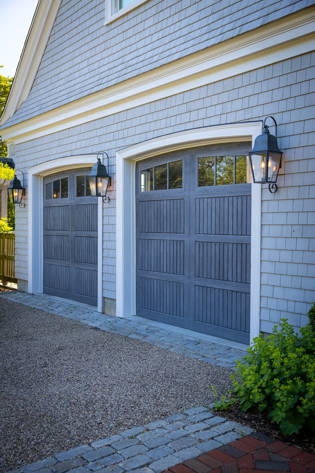 A driveway with a textured gravel surface leading to a double garage with blue paneled doors. Attached to the house exterior are two black lantern-style outdoor lights, and there is greenery on the right side of the image.
