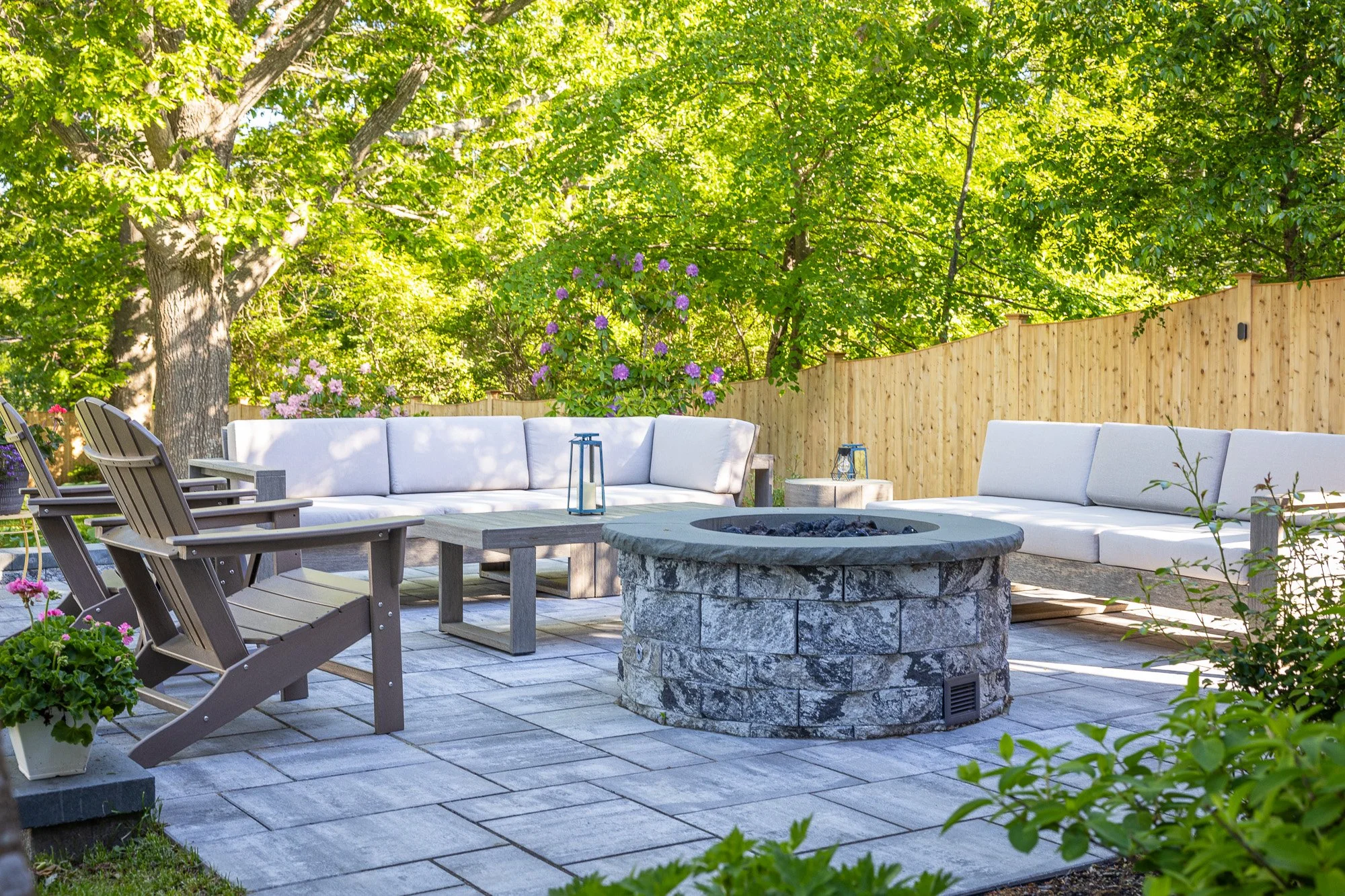 An outdoor patio with a stone fire pit, white cushioned seating, wooden chairs, and a view of lush green trees and flowering plants behind a wooden fence.