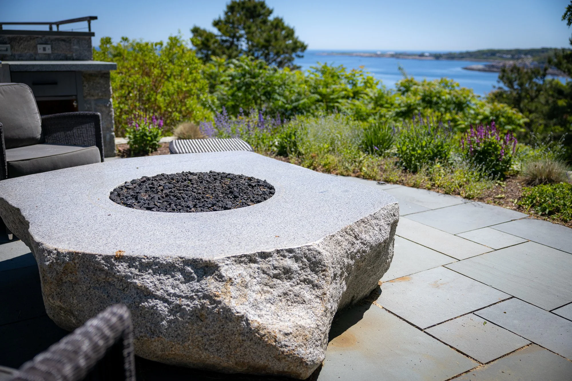 Outdoor patio area with a stone fire pit, a cushioned chair, and a view of trees, water, and distant land under a clear blue sky.