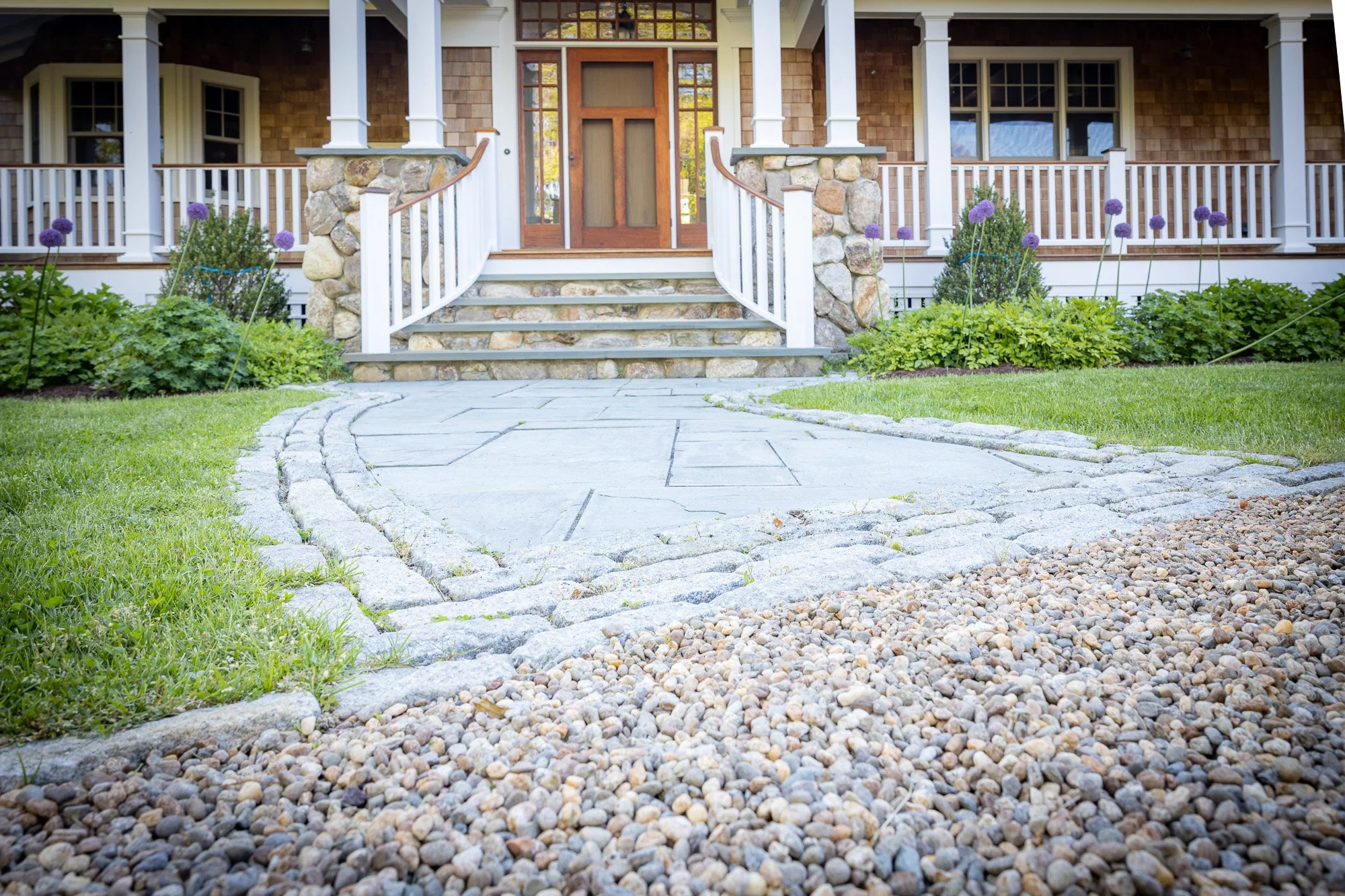 Front porch and entryway of a house with stairs, stone foundation, white railings, wooden siding, and purple flowers in garden beds.
