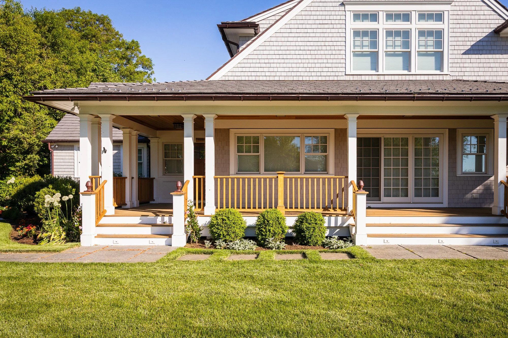 Front view of a two-story house with a porch, white columns, a wooden railing, and exterior windows, surrounded by a green lawn and trees under a clear blue sky.