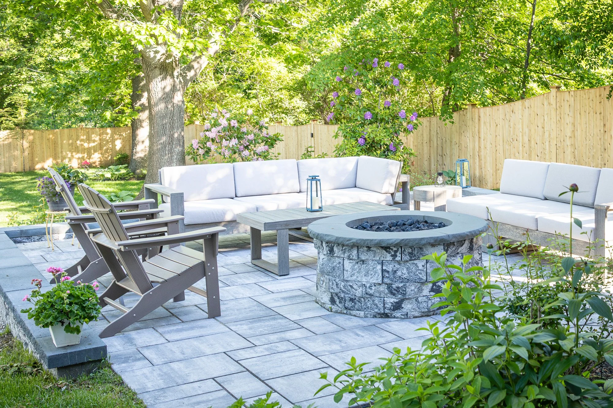 A backyard patio scene with white cushioned outdoor furniture, potted plants, a fire pit, and a wooden privacy fence, surrounded by lush green trees and plants.
