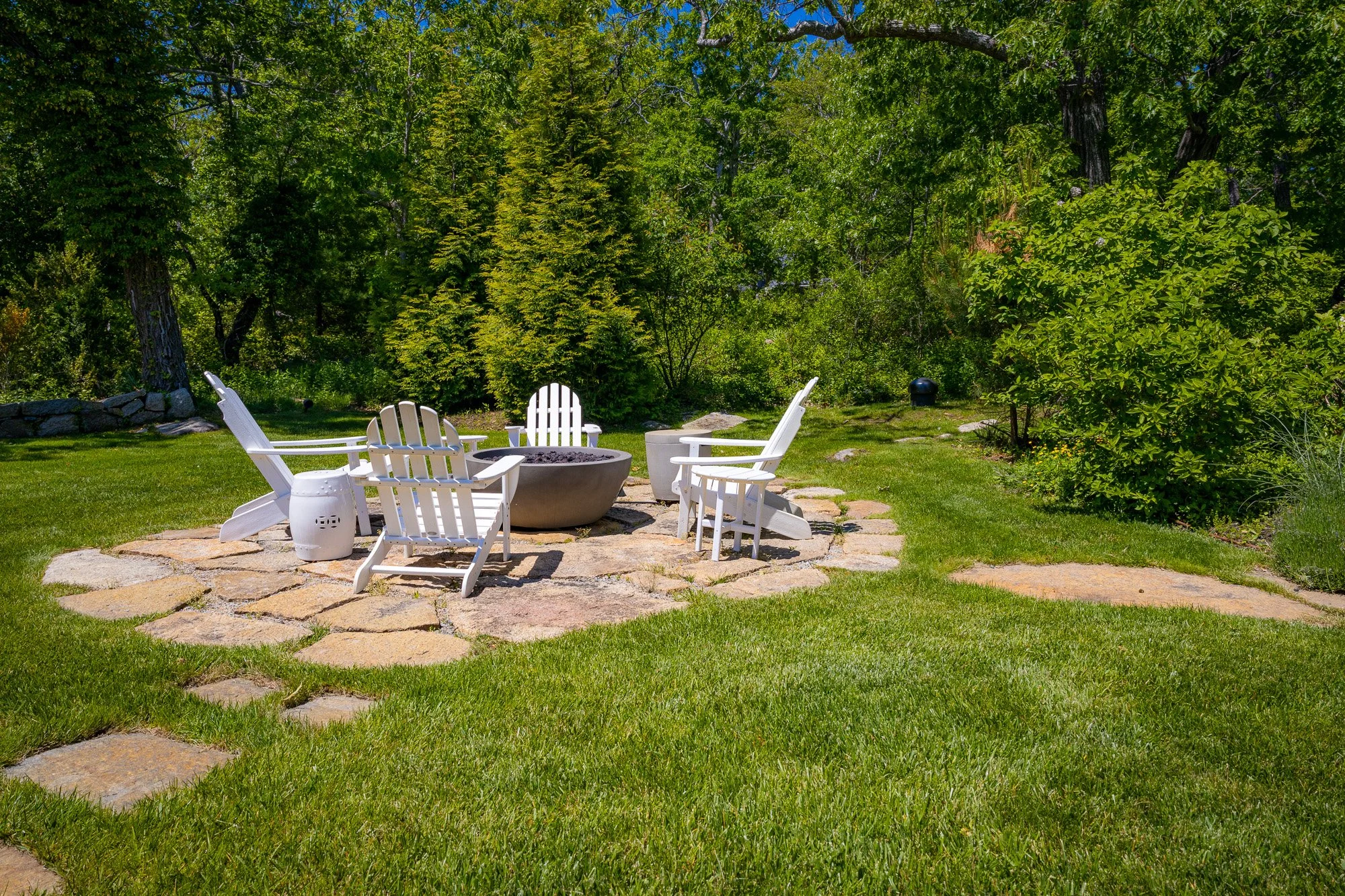 A backyard garden with four white Adirondack chairs arranged around a fire pit on a stone patio. The area is surrounded by lush green grass and tall trees with dense foliage under a clear blue sky.