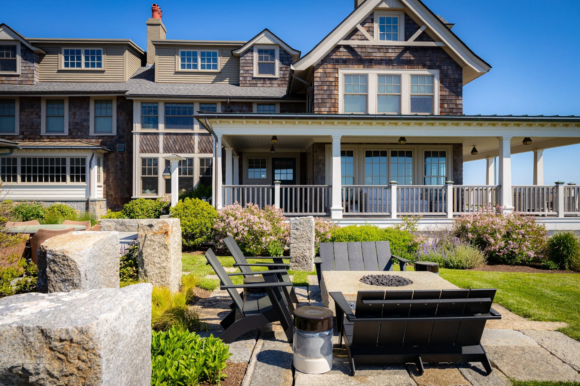 Backyard patio with black Adirondack chairs around a fire pit, surrounded by rocks, shrubs, and pink flowers, with a large house in the background.