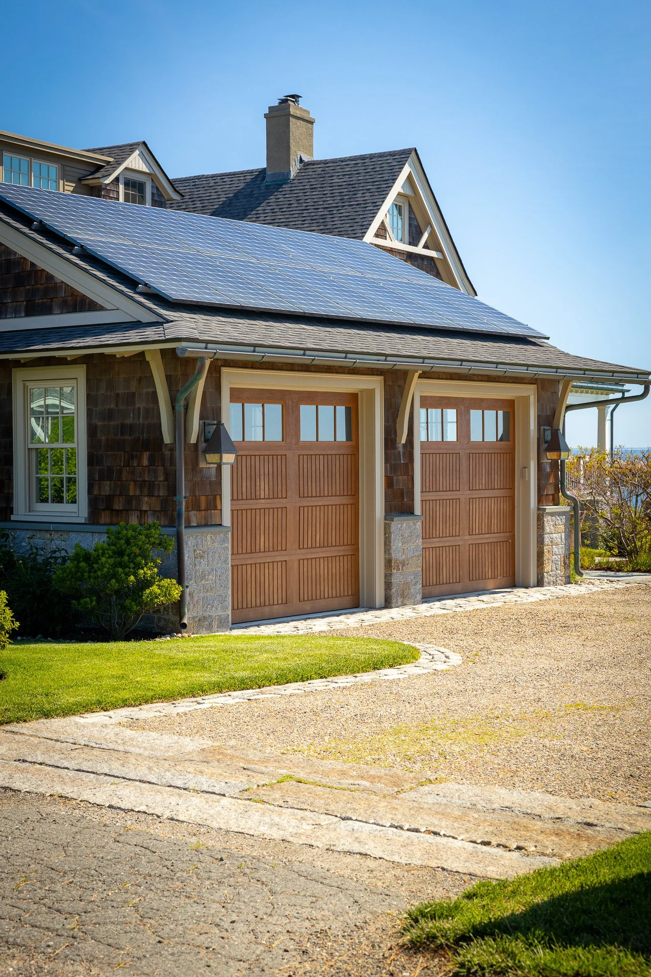 A house with solar panels on the roof, a two-car garage with wooden doors, a well-maintained lawn, and a gravel driveway.